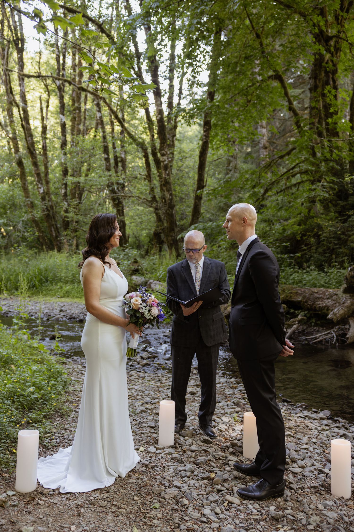 Intimate outdoor wedding ceremony taking place by a creek in a wooded forest setting with white candle decorations.