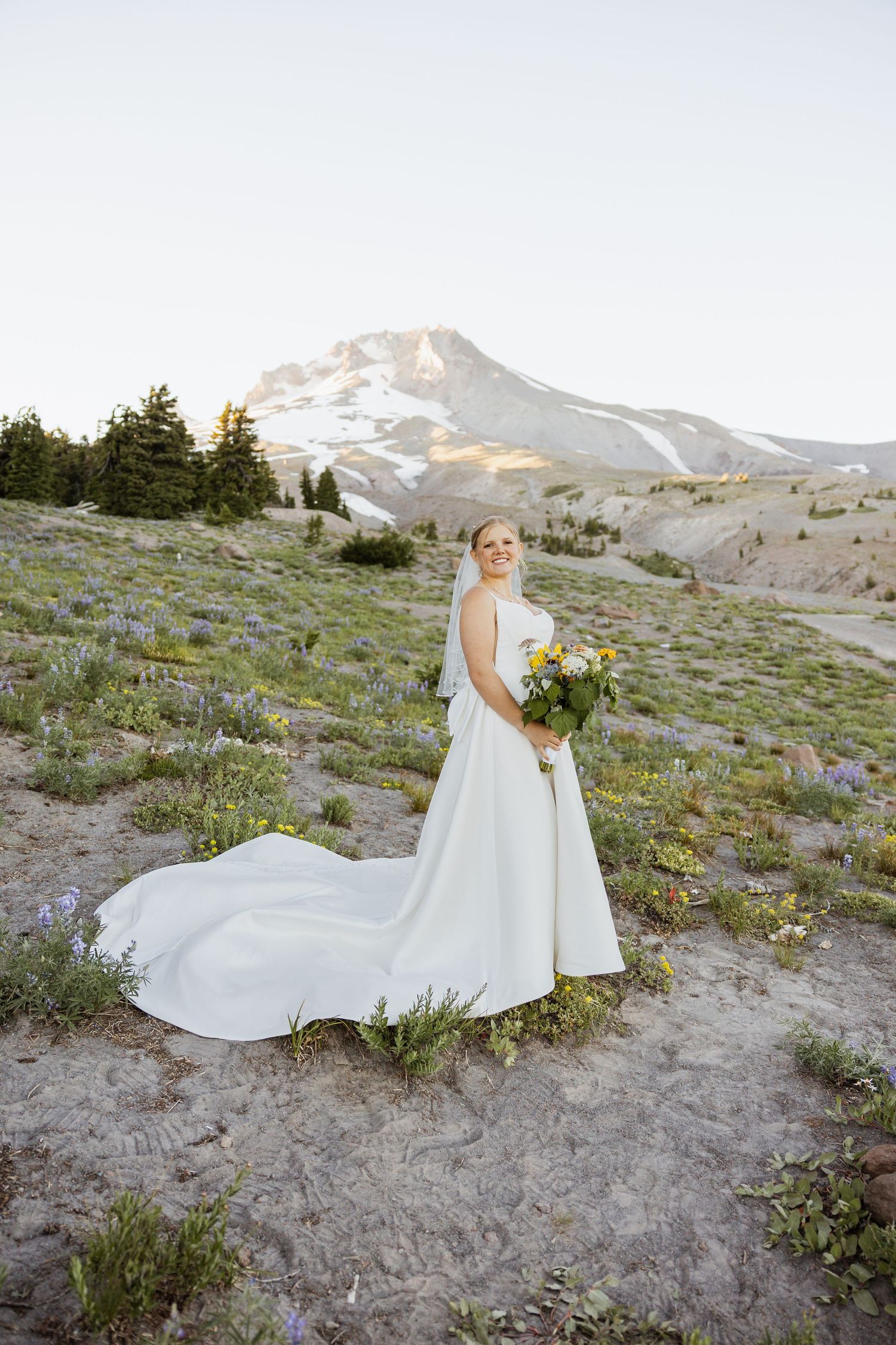 A white wedding dress flows across mountain wildflowers with snow-capped peaks in the background.