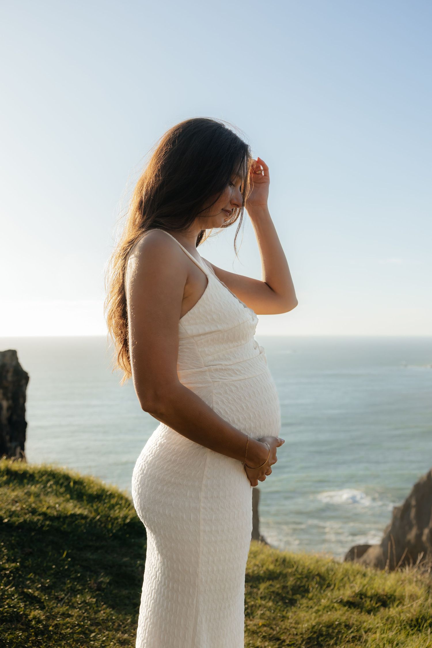 Pregnant woman in white dress holding belly by ocean cliffs on sunny day.