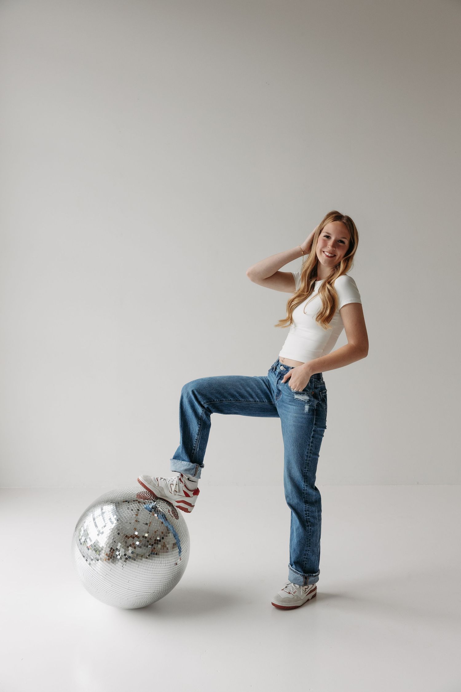 Young woman in jeans and white top posing with a large silver disco ball in a bright studio setting.