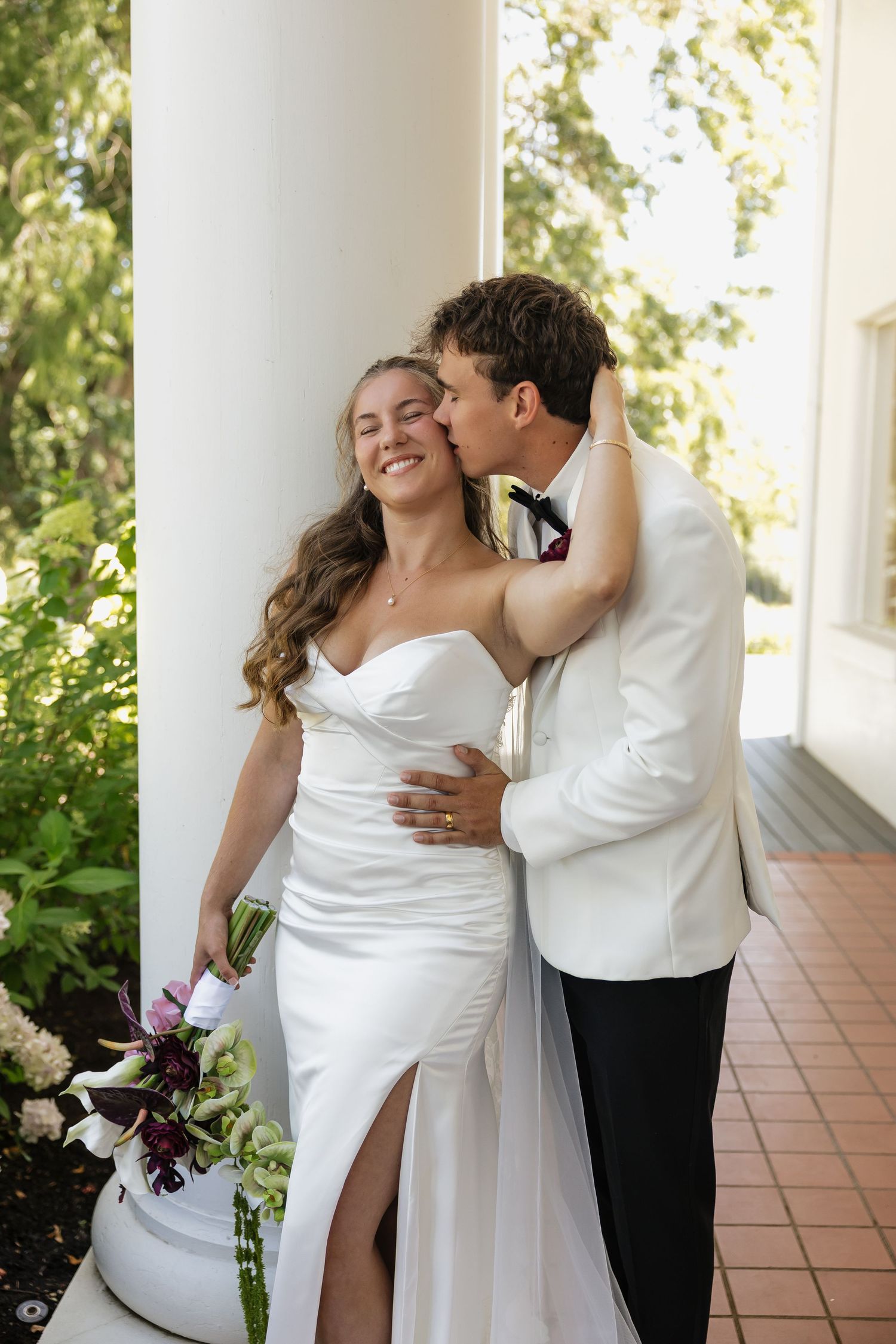 A groom in a white suit kisses his smiling bride on the cheek as she holds a bouquet of dark florals.
