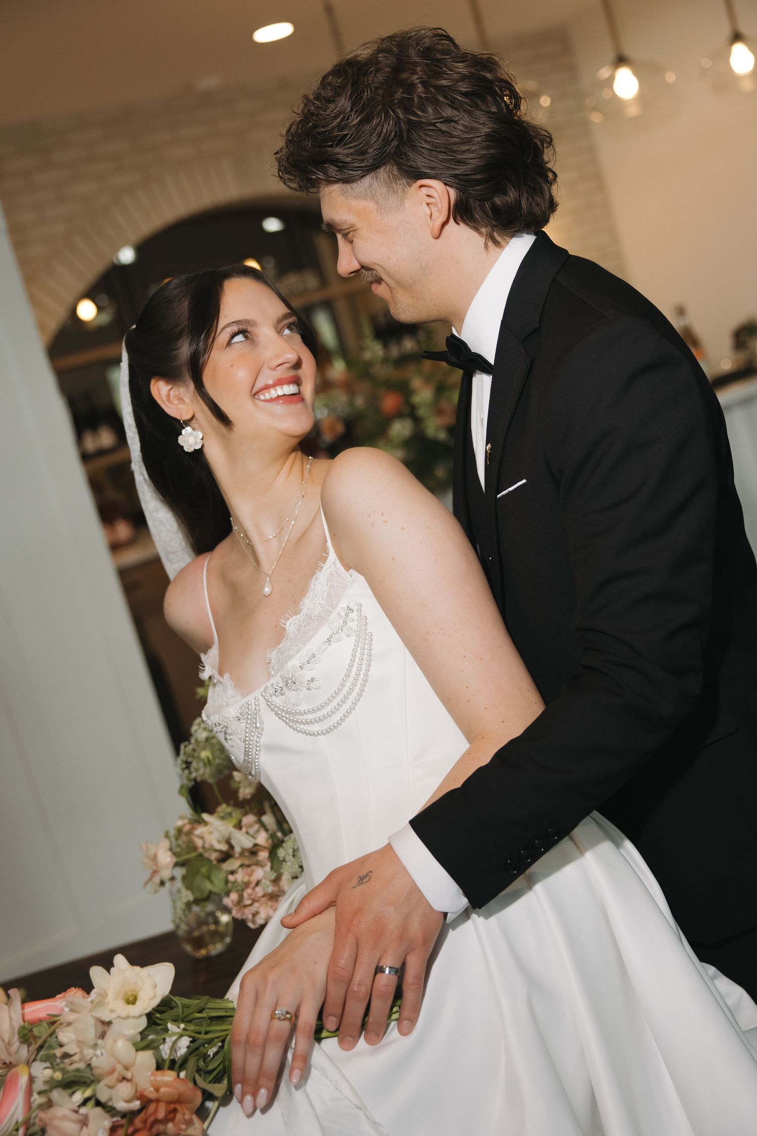 Smiling bride in white gown with veil and groom in black tuxedo sharing a joyful moment at their wedding reception.