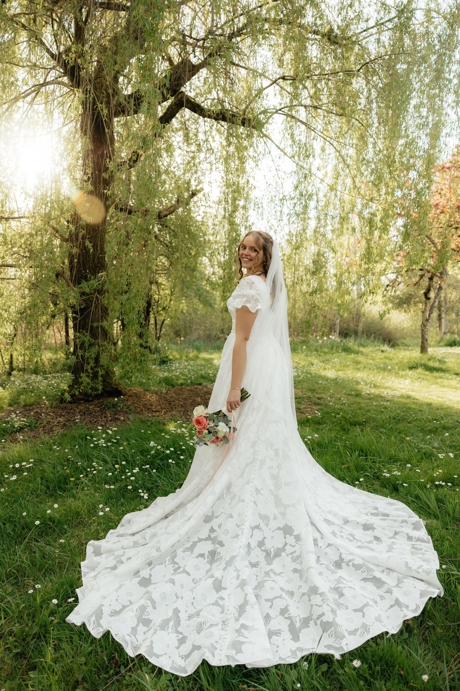 Smiling bride in lace wedding gown with long train and veil holding bouquet under weeping willow tree.