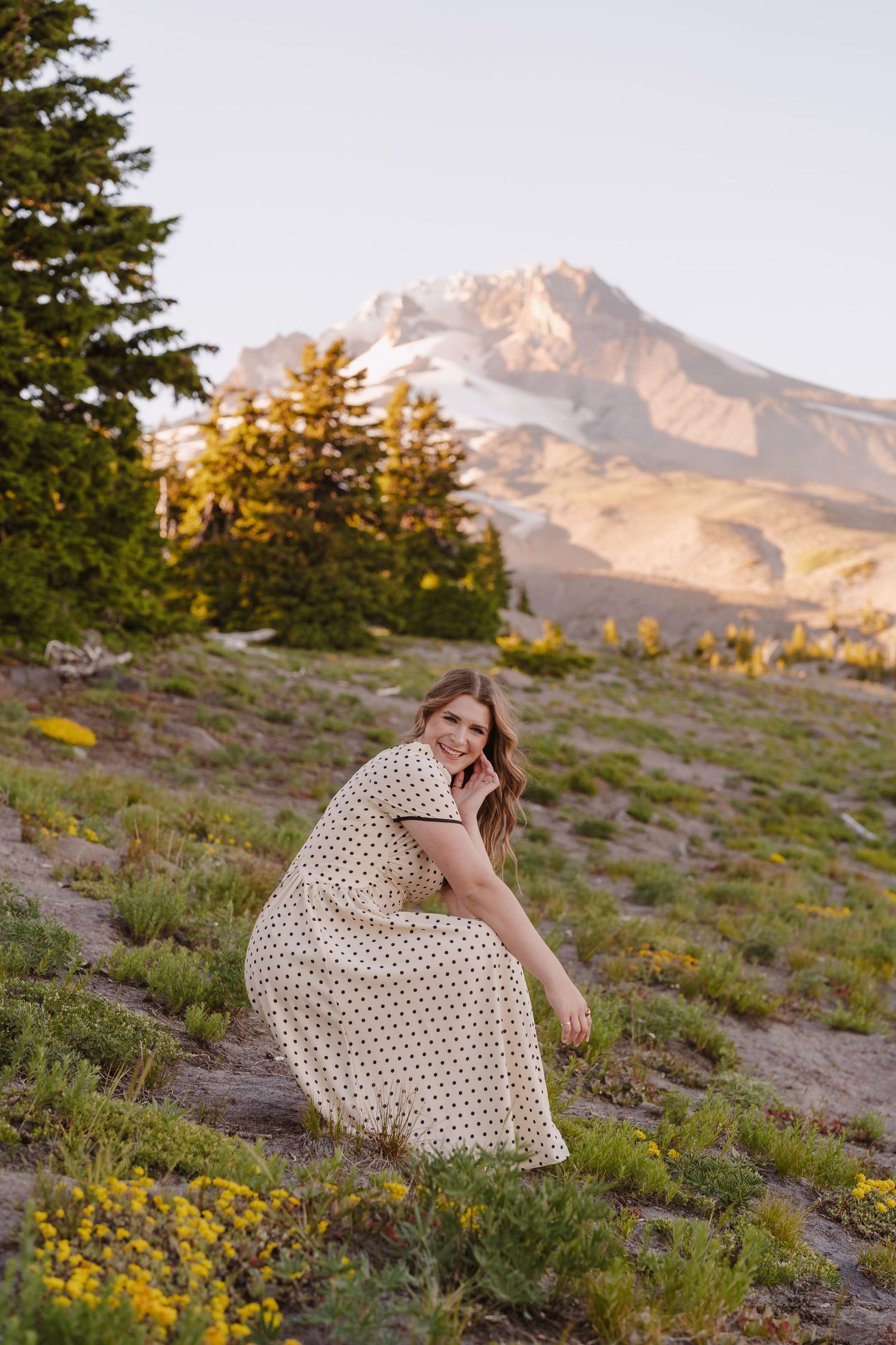 Woman in polka dot dress crouching on alpine meadow with Mount Hood and evergreen trees in background at sunset.