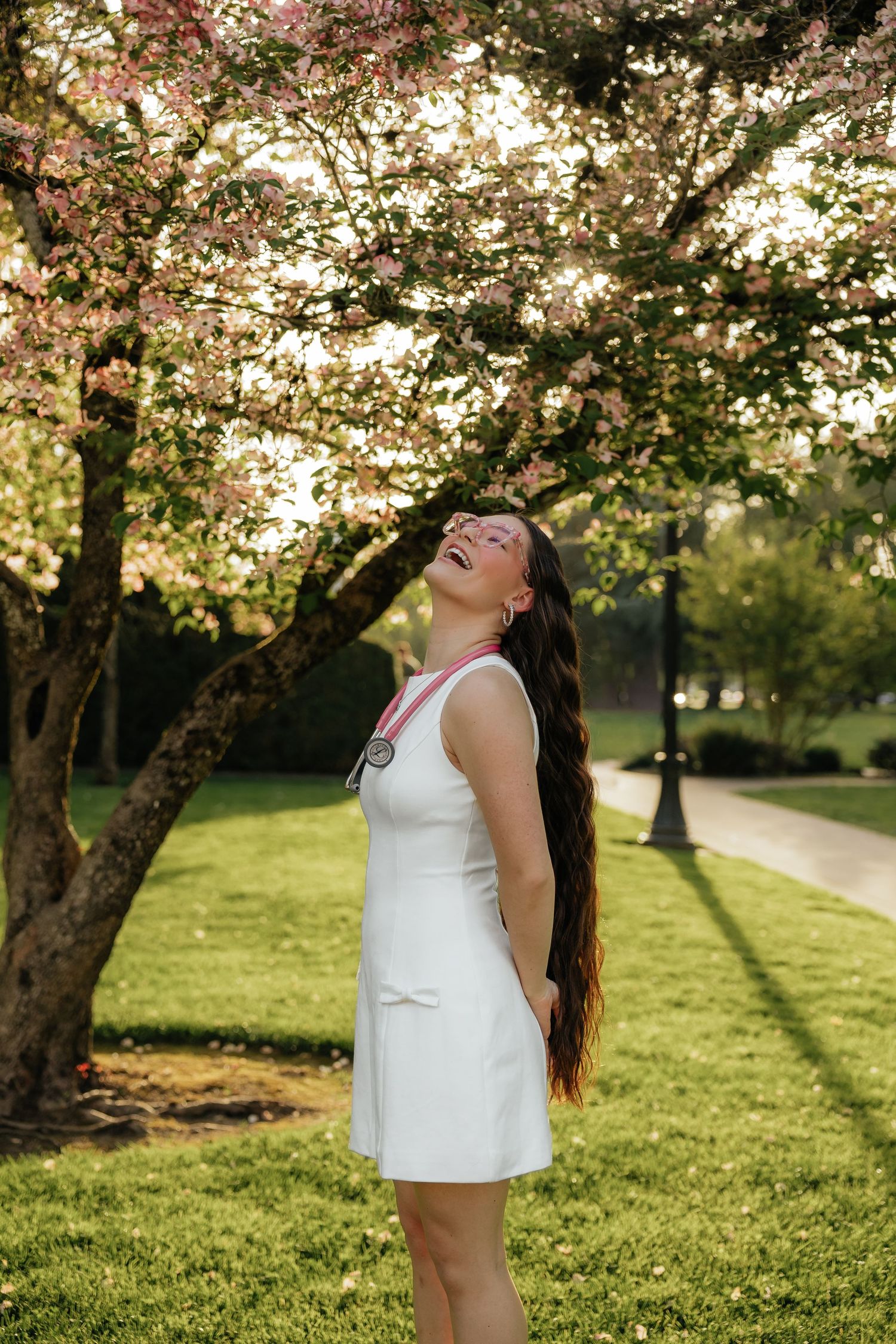 Young woman in white dress laughing under pink cherry blossom tree in sunny park.