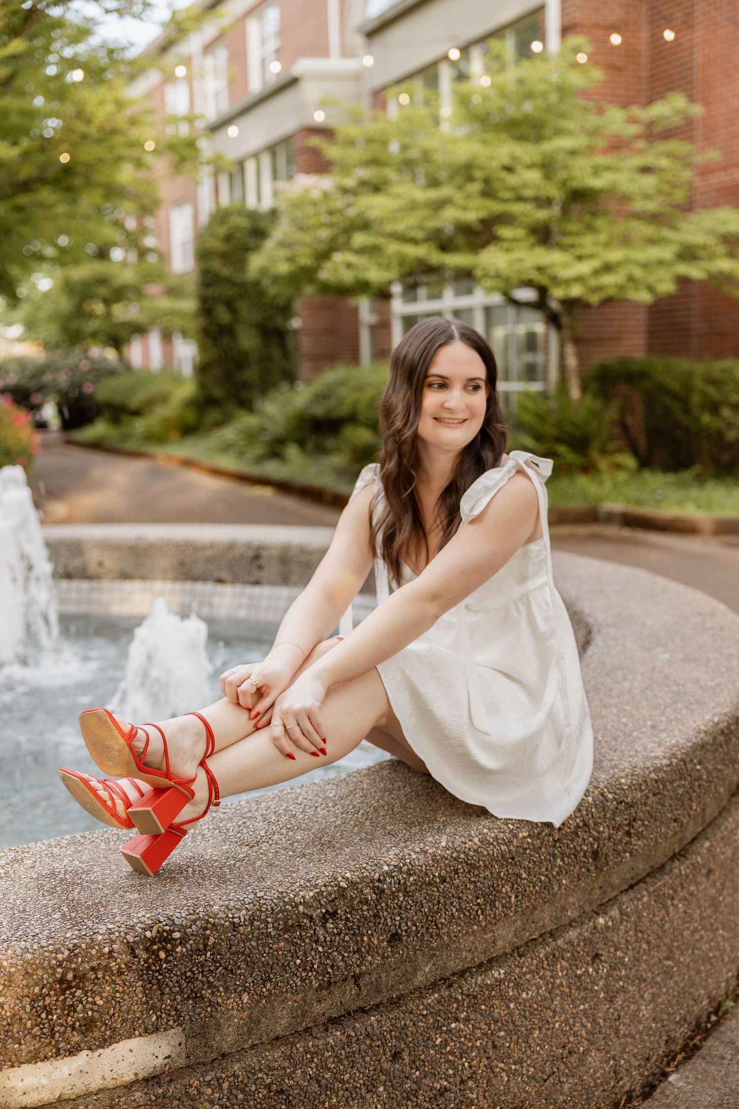 A person in a white dress adjusts their bright red high heels while sitting on a concrete wall outdoors.