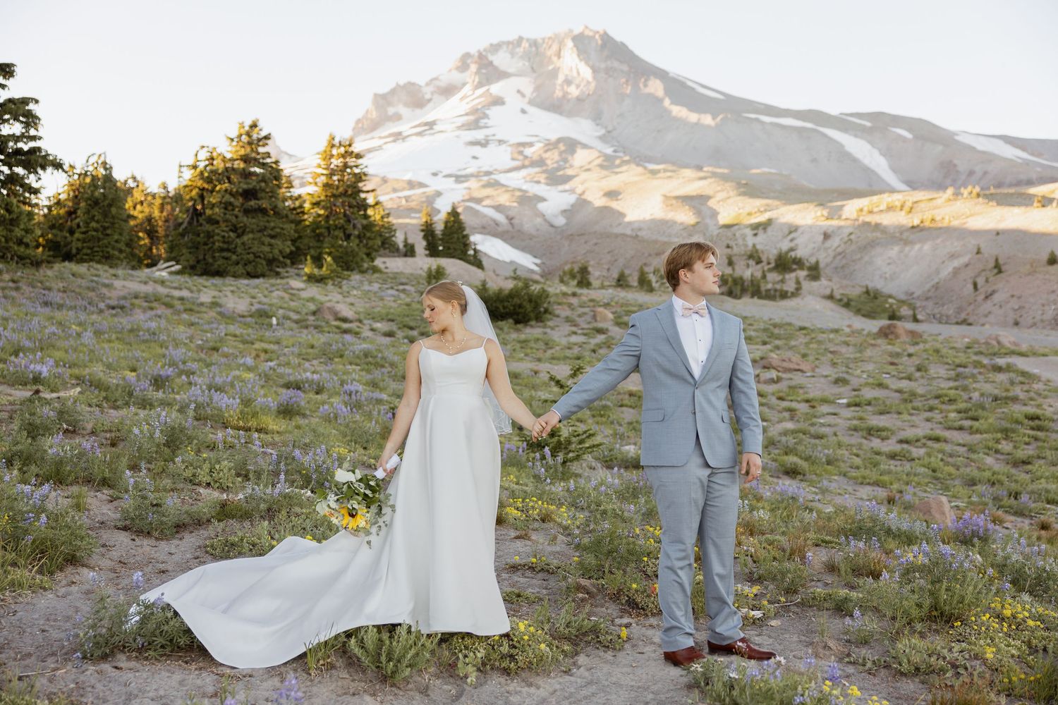A couple in wedding attire stands in a mountain meadow at sunset with snow-capped peaks in the distance.