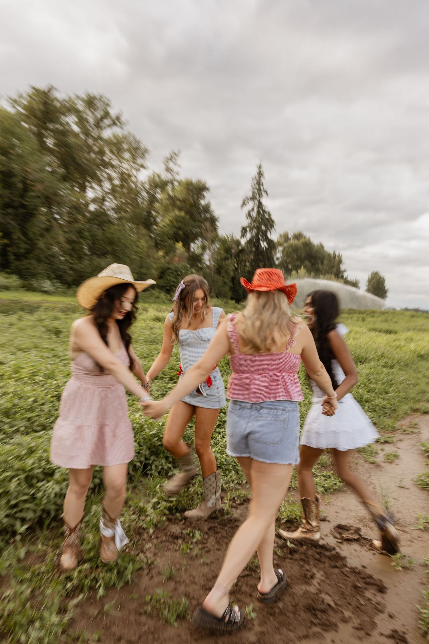 Four friends in summer outfits walk together on a dirt path through green fields under cloudy skies.