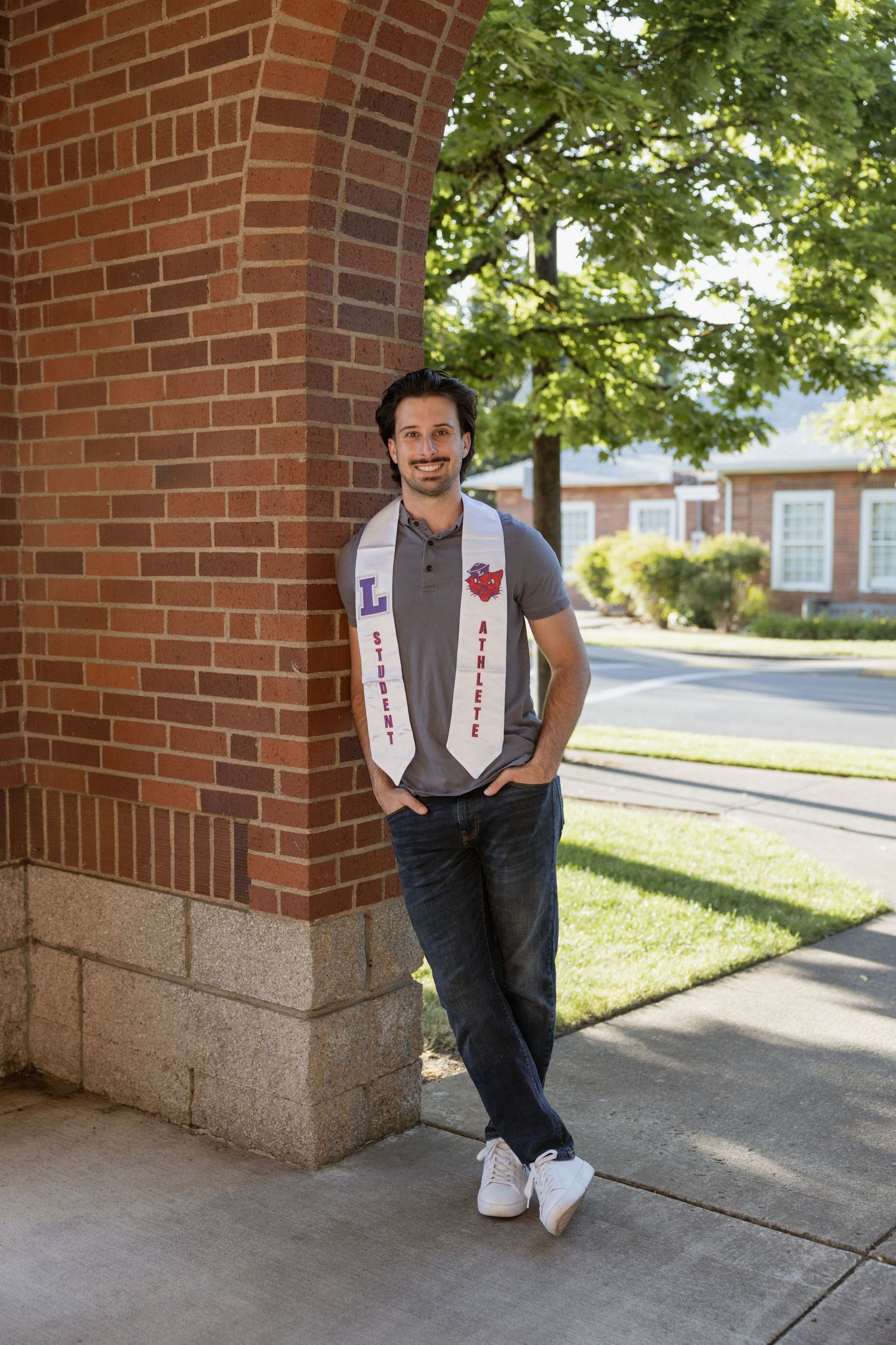 A student in a gray vest and black pants poses against a brick wall in a series of casual graduation photos.