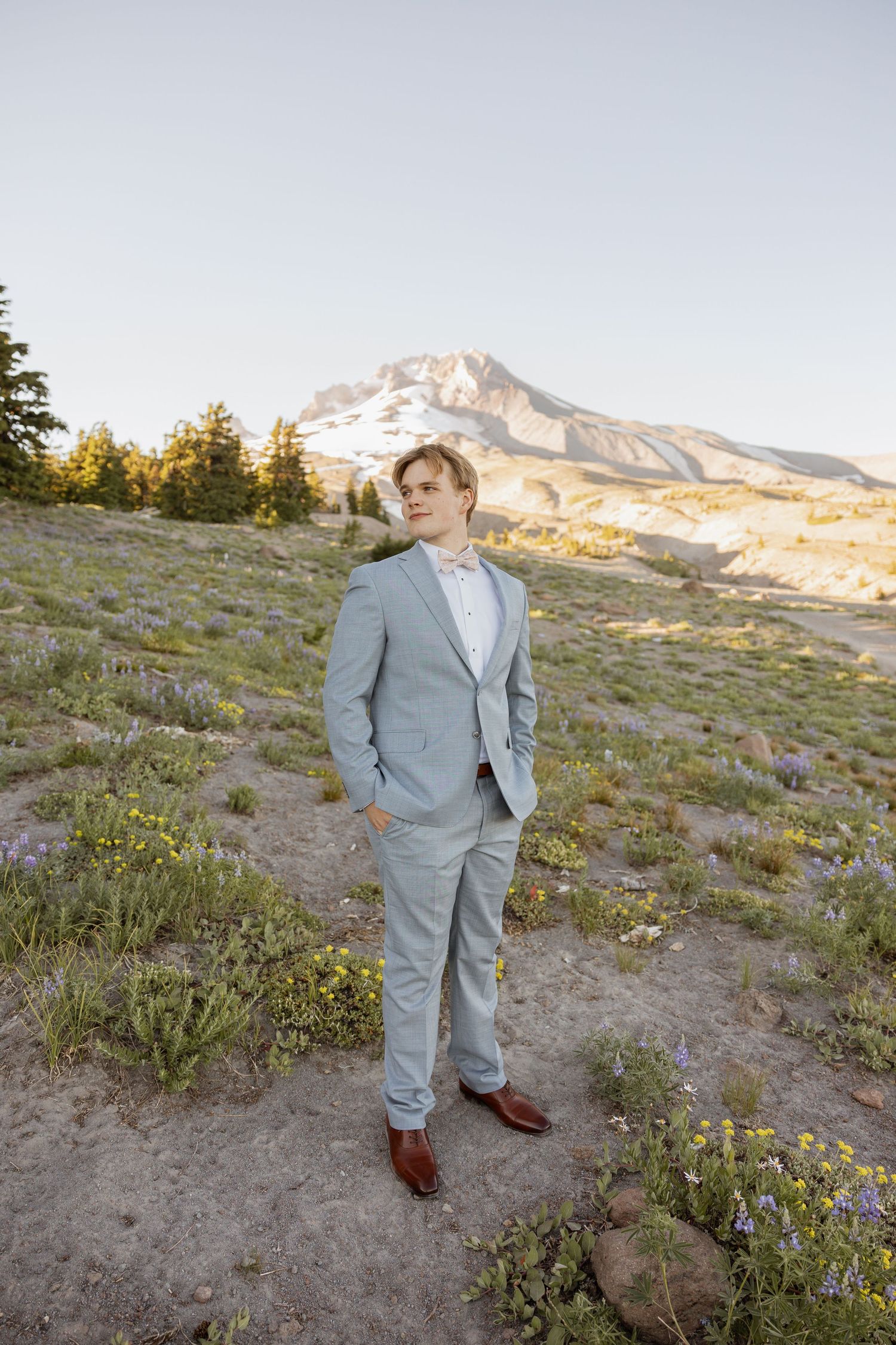 A person in a light gray suit stands on a mountain slope with wildflowers and snow-capped peaks in the background.