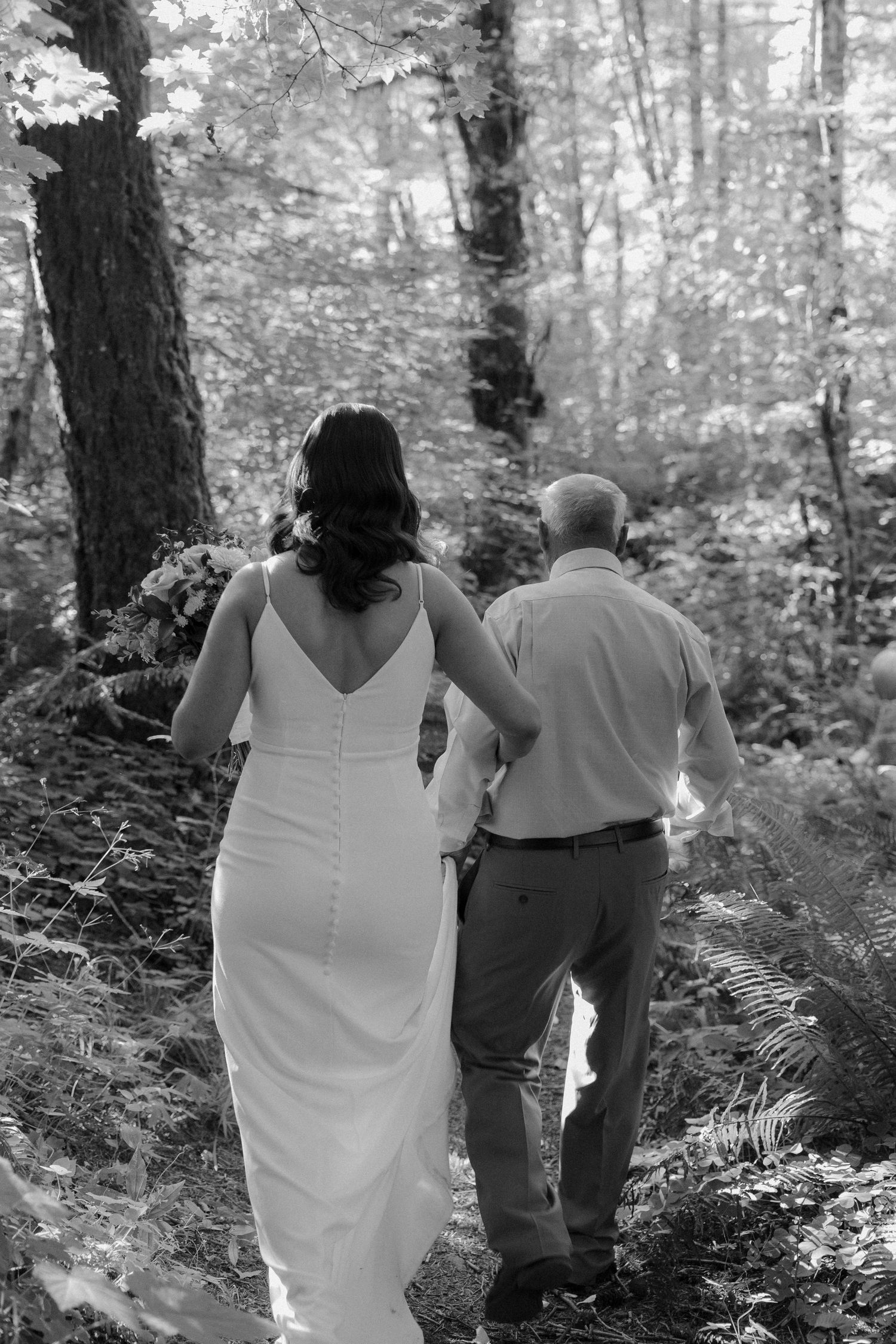 Black and white photo of two people walking together through a wooded forest path on a sunny day.
