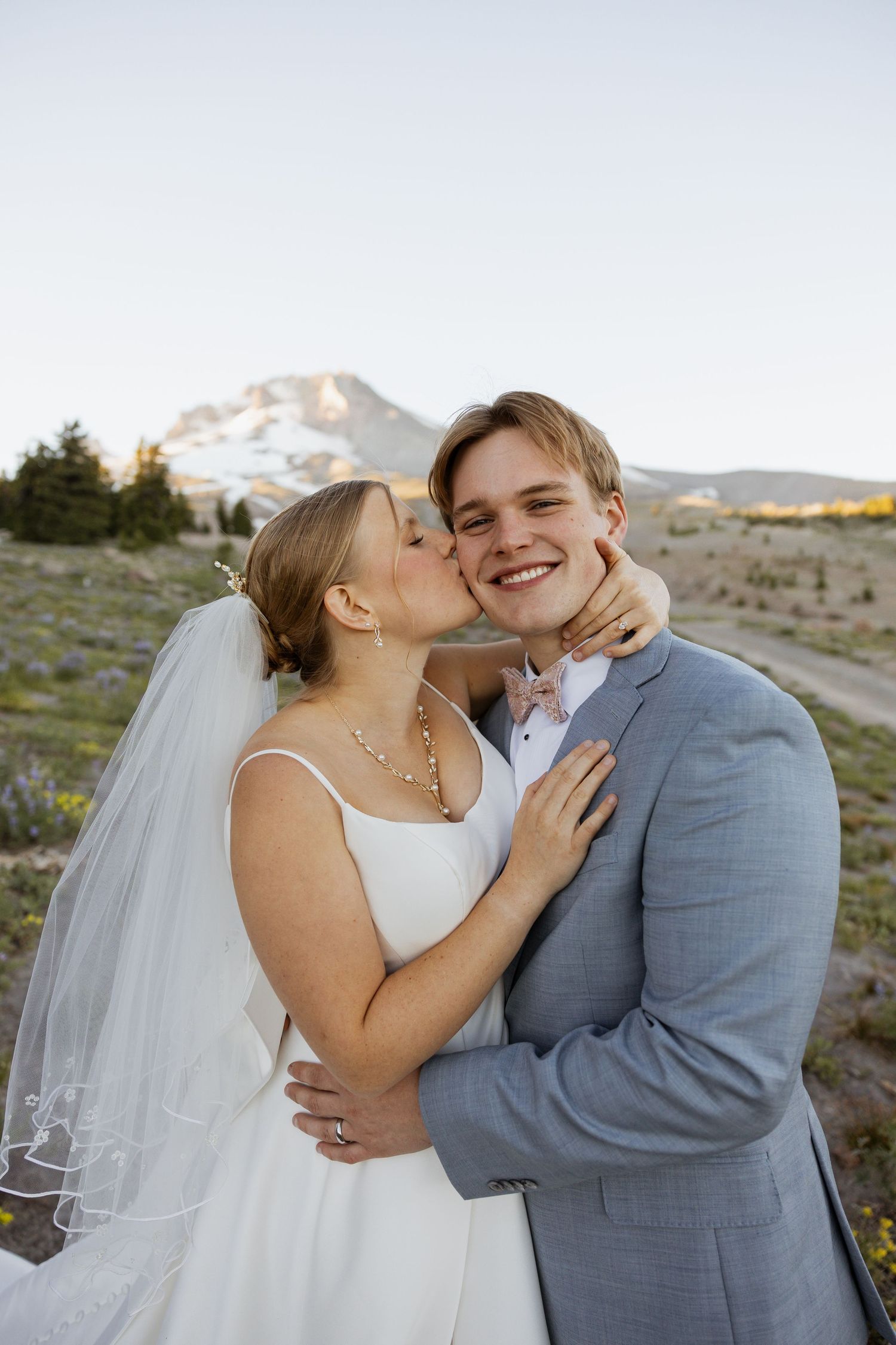 A joyful wedding couple embraces and laughs together against a scenic mountain backdrop at sunset.