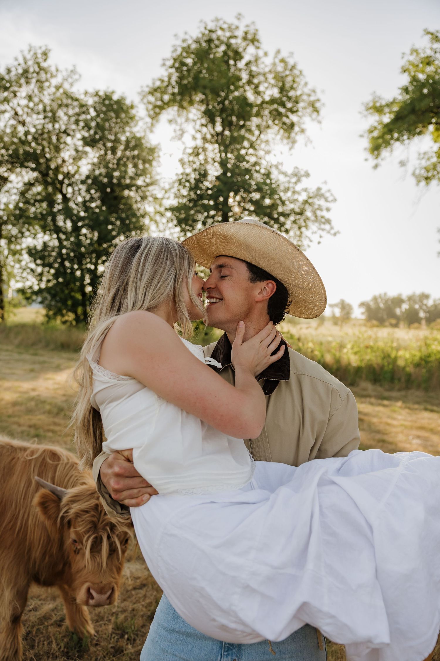 Couple sharing a romantic kiss outdoors on a farm, man in cowboy hat holding woman in white dress with Highland cow nearby.