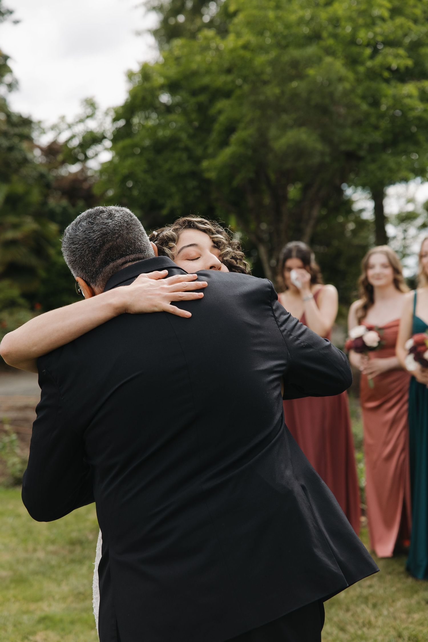 Bride and groom share emotional hug at outdoor wedding ceremony while bridesmaids in burgundy and dusty rose dresses look on.