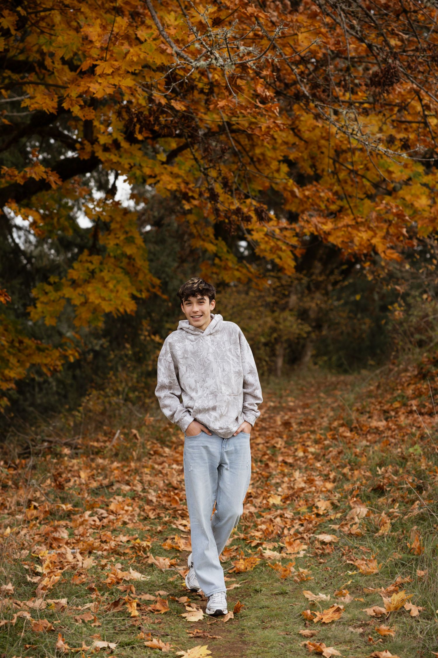 Someone in a cozy beige hoodie and light jeans takes an autumn stroll beneath golden maple trees on a leaf-strewn path.
