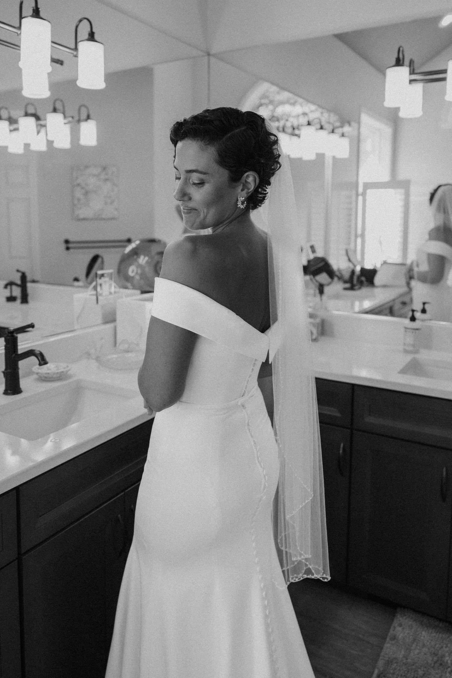 Black and white photograph captures bride's elegant dress details in bathroom mirror reflection.