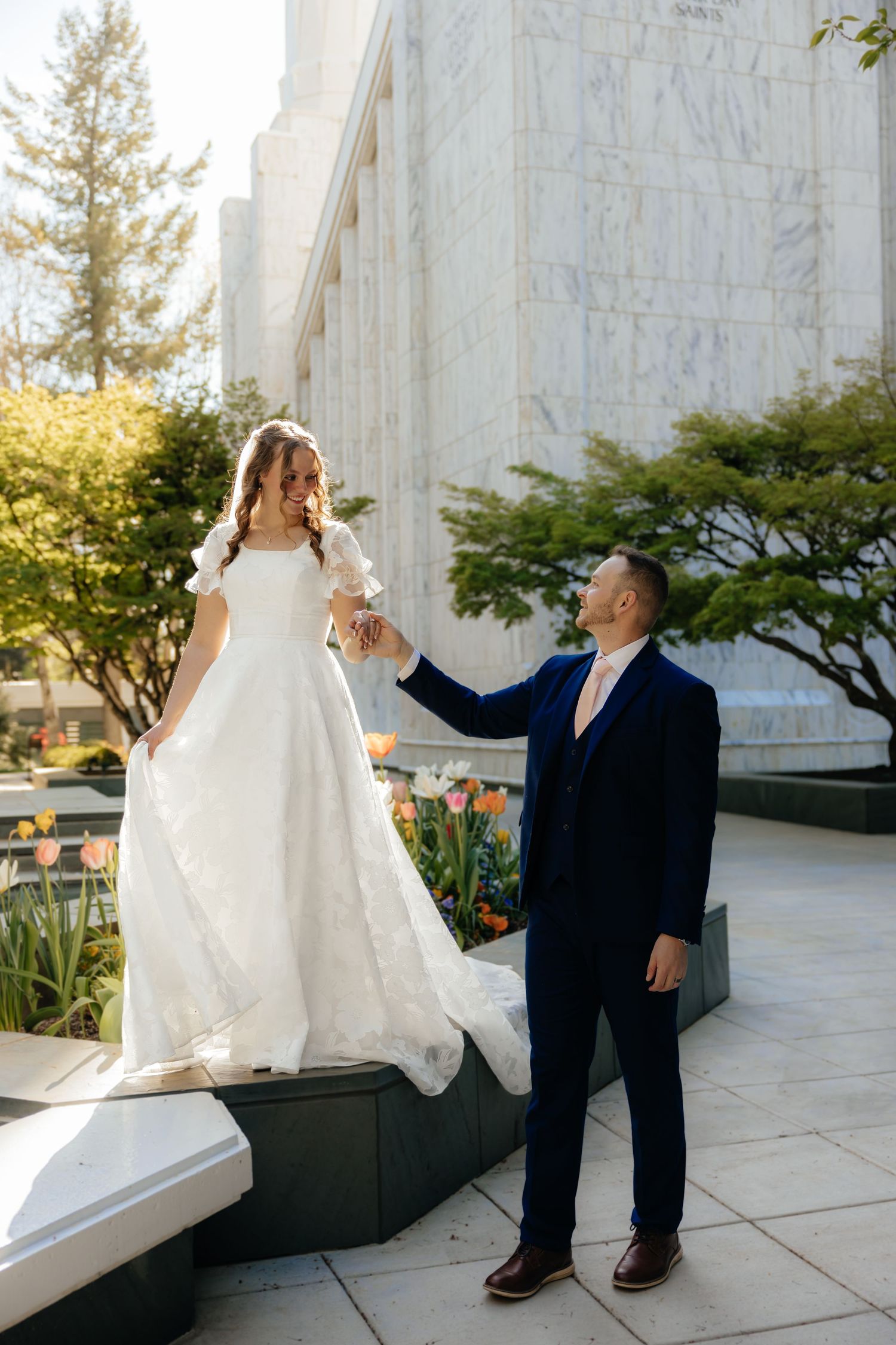 Bride in white gown stands elevated as groom in navy suit holds her hand outside a marble temple.