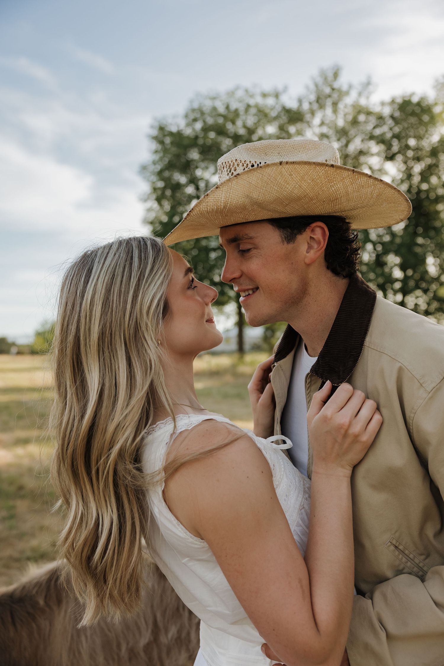 A couple shares a romantic moment outdoors, the man wearing a straw cowboy hat and tan jacket, the woman in a white dress.