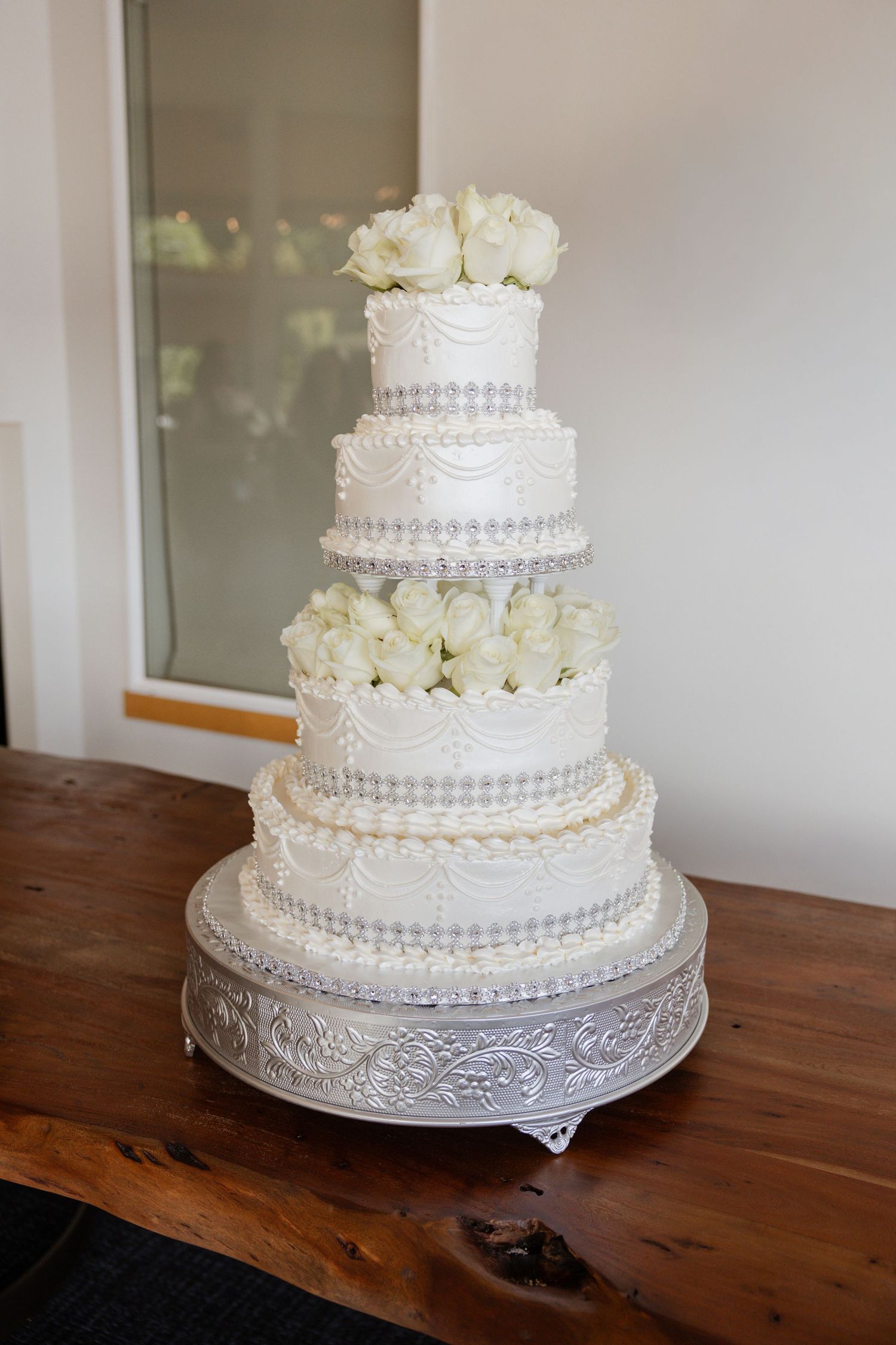 Elegant white wedding cake with four tiers decorated with white roses and pearl details on a wooden table.