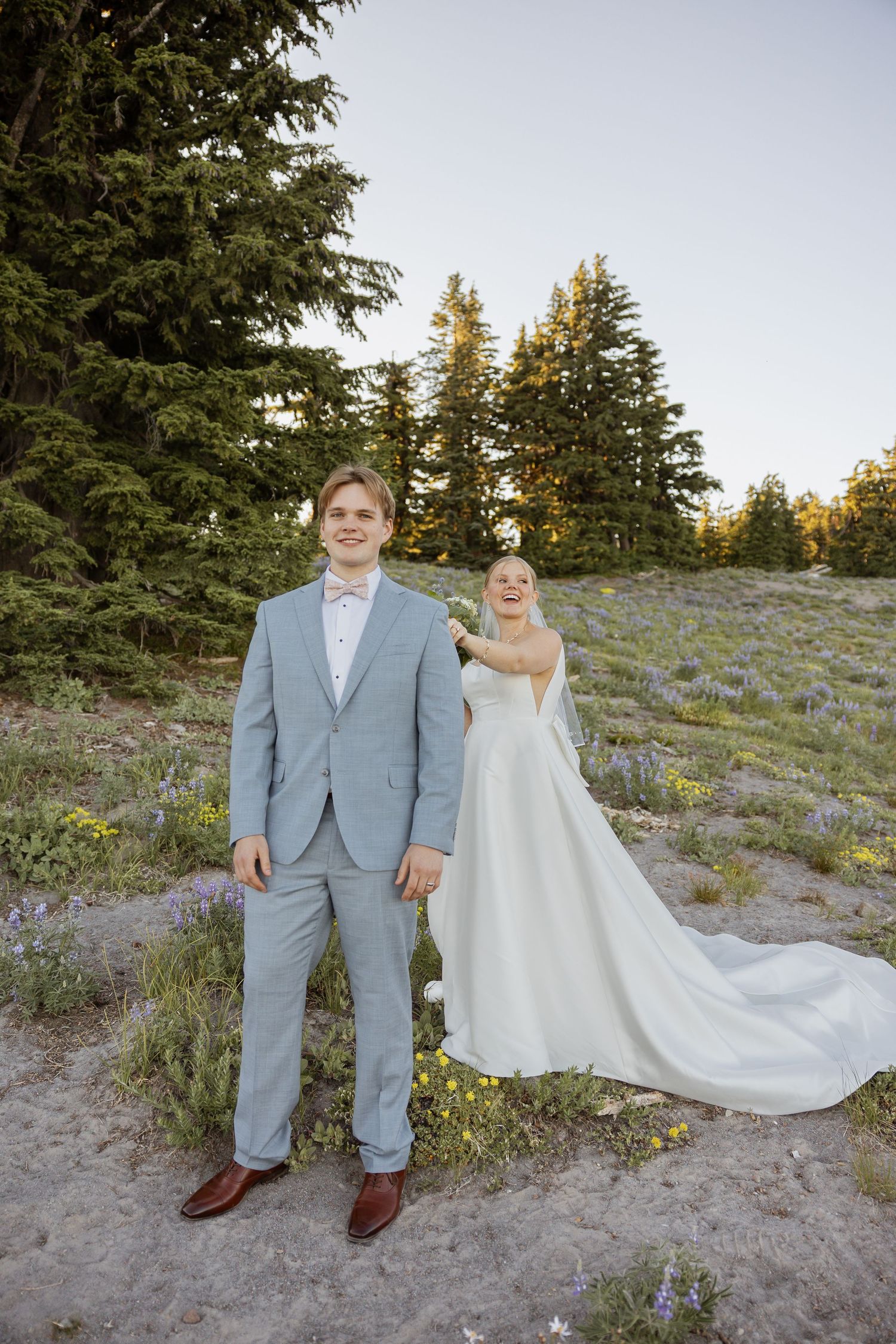 A wedding couple poses on a mountainside with evergreen trees and wildflowers during golden hour.