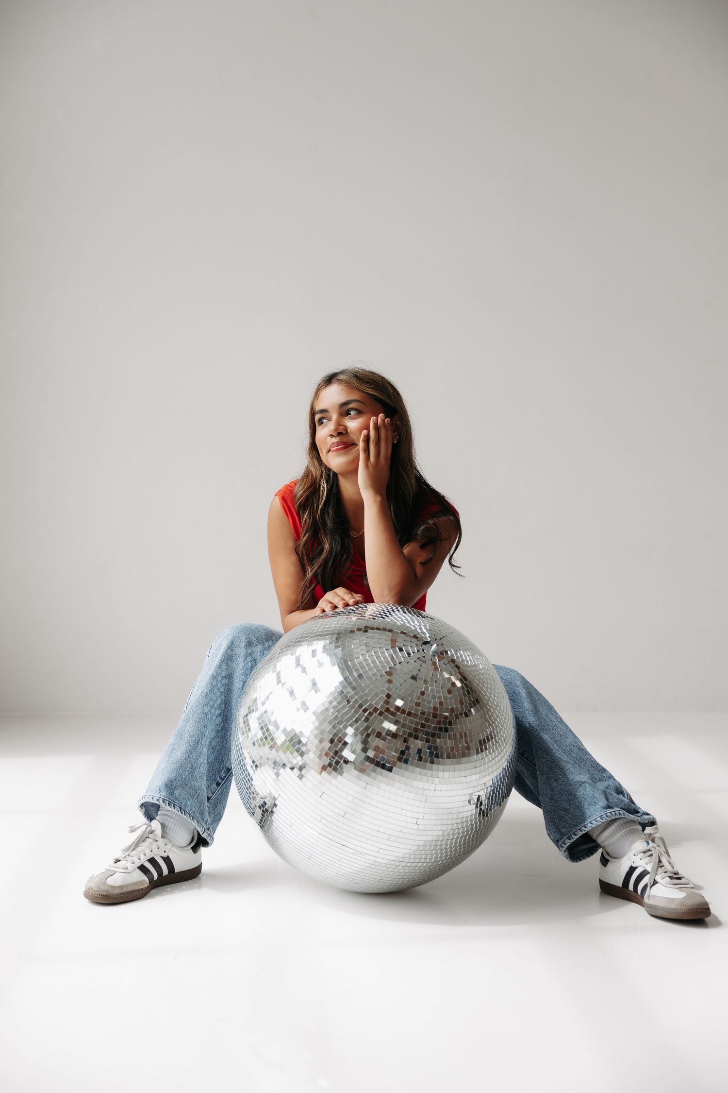 Young woman in red top and jeans sitting on floor with large silver disco ball in white studio.