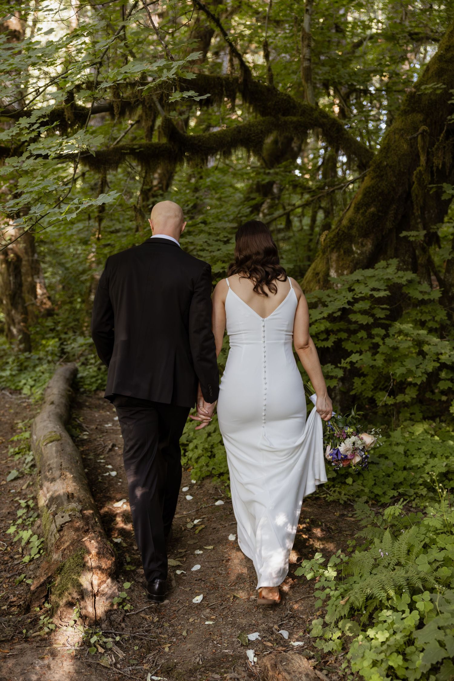 A couple in wedding attire walks down a wooded forest path surrounded by lush green foliage.