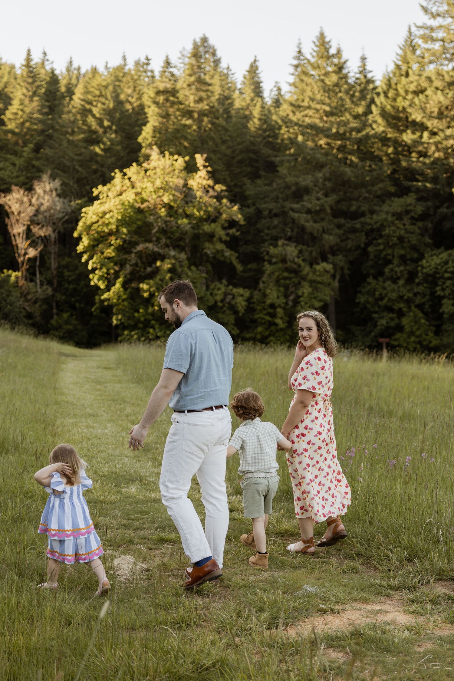 A group walks together along a grassy trail through evergreen forest on a sunny summer evening.