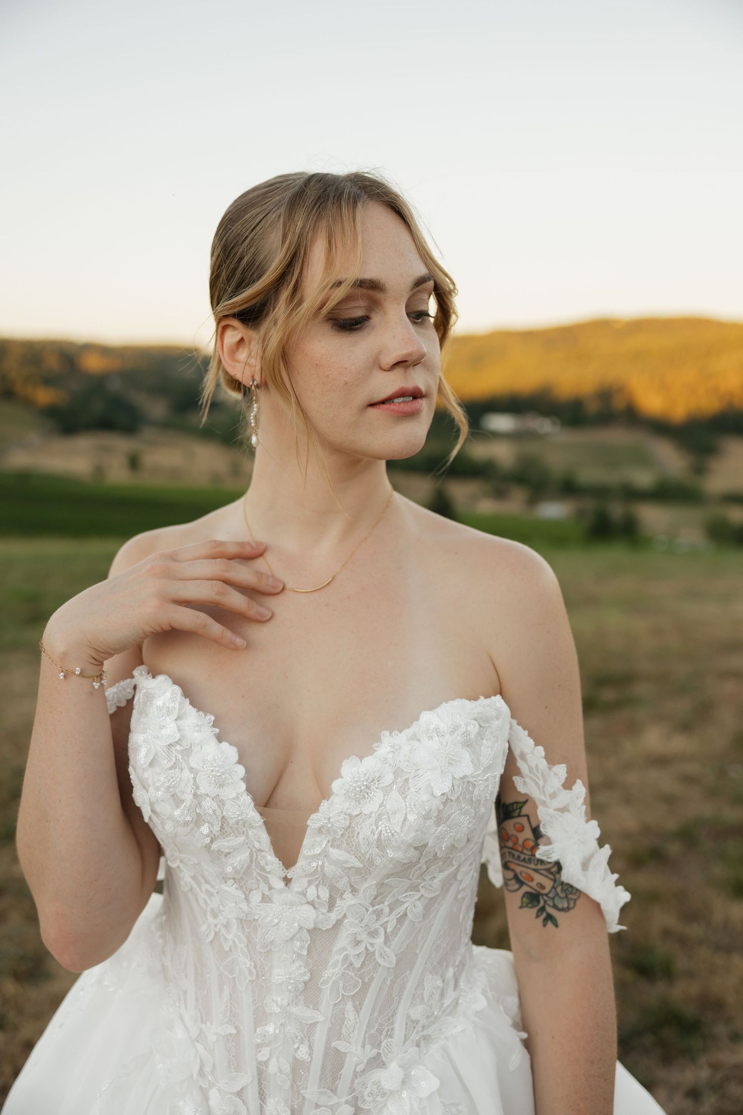 Bride in off-shoulder lace wedding gown with floral appliques poses outdoors at golden hour with rolling hills.