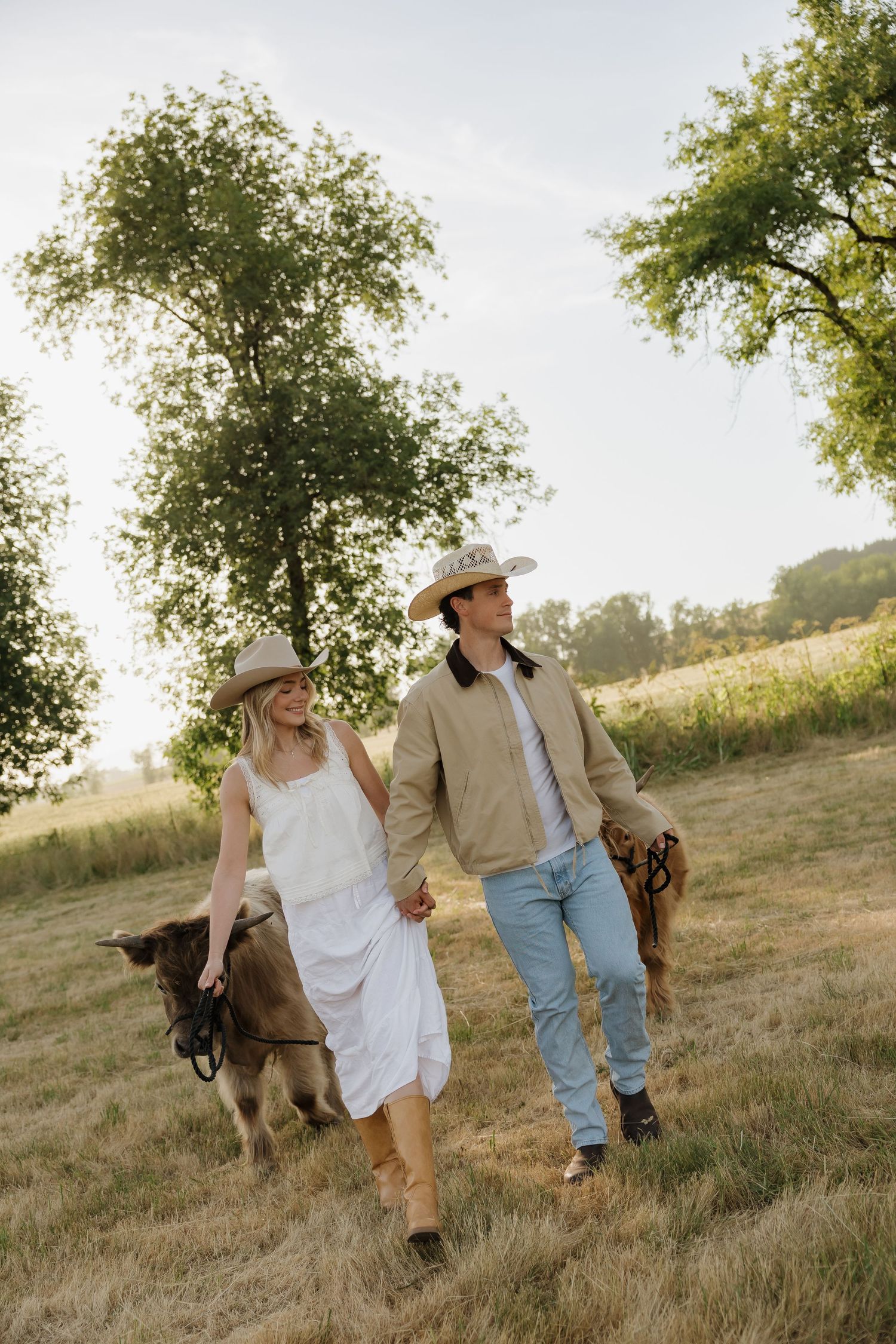 Couple in cowboy hats walking hand in hand through a sunny field with a small cow and trees in background.