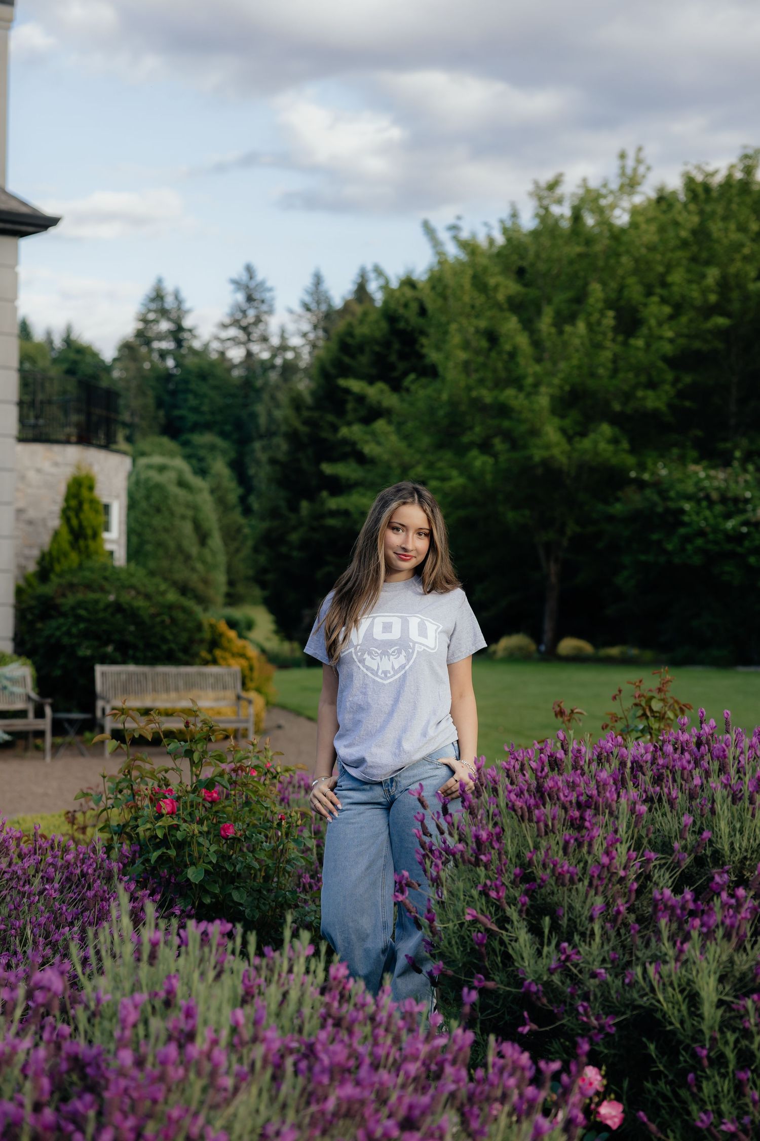 Young woman in gray VDU t-shirt and jeans standing among purple lavender flowers in a lush garden.