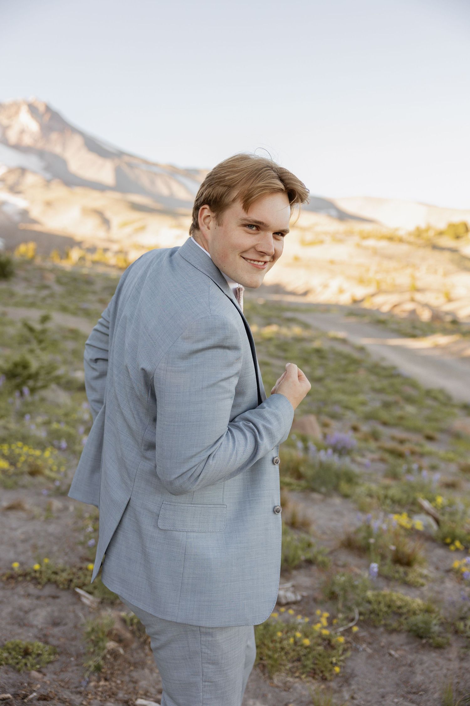 Someone in a light gray suit poses outdoors against a mountainous desert landscape at golden hour.