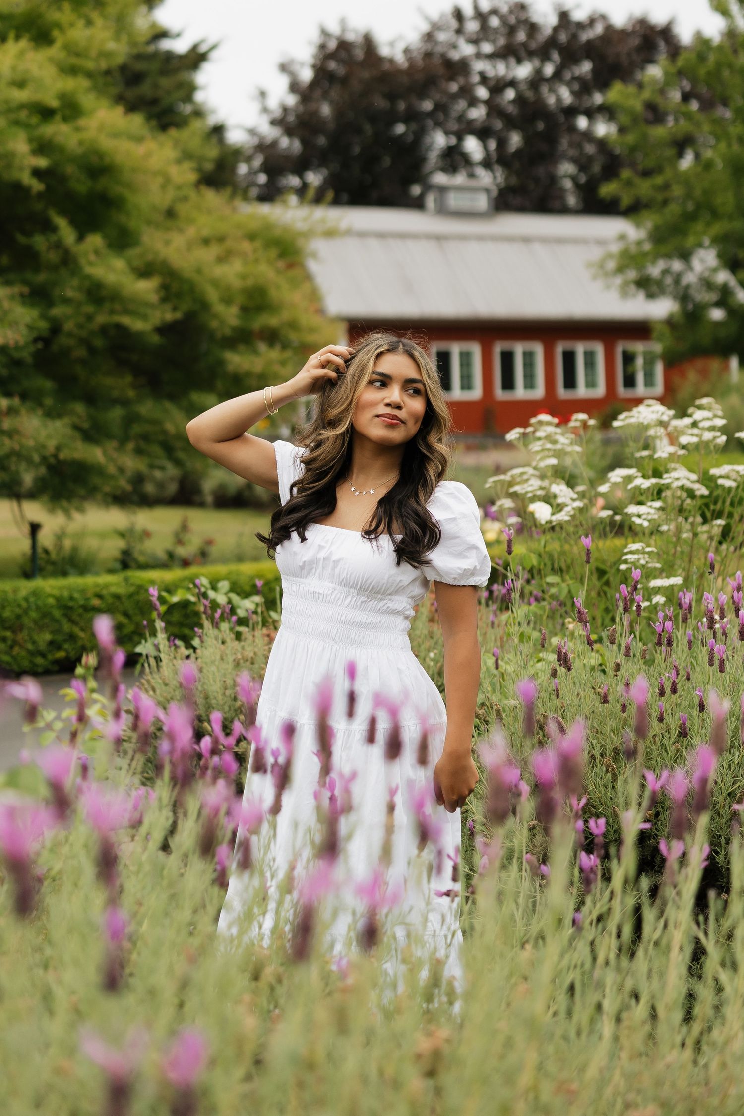 Young woman in white puff-sleeve dress standing in lavender garden with red barn in background.