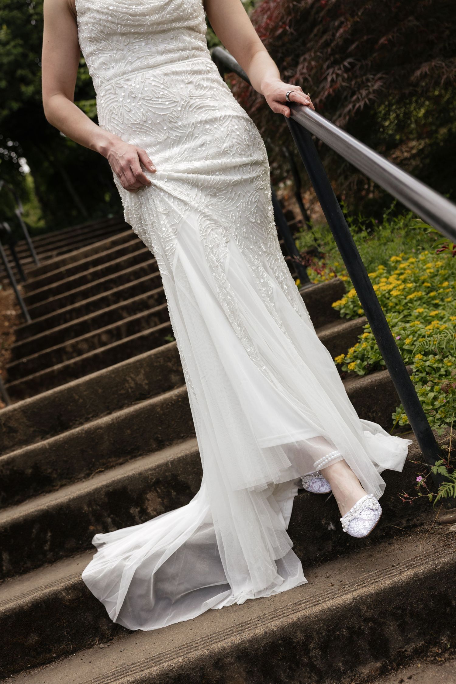 Bride in beaded white wedding gown with train walking up stone steps, holding railing, wearing white lace shoes.