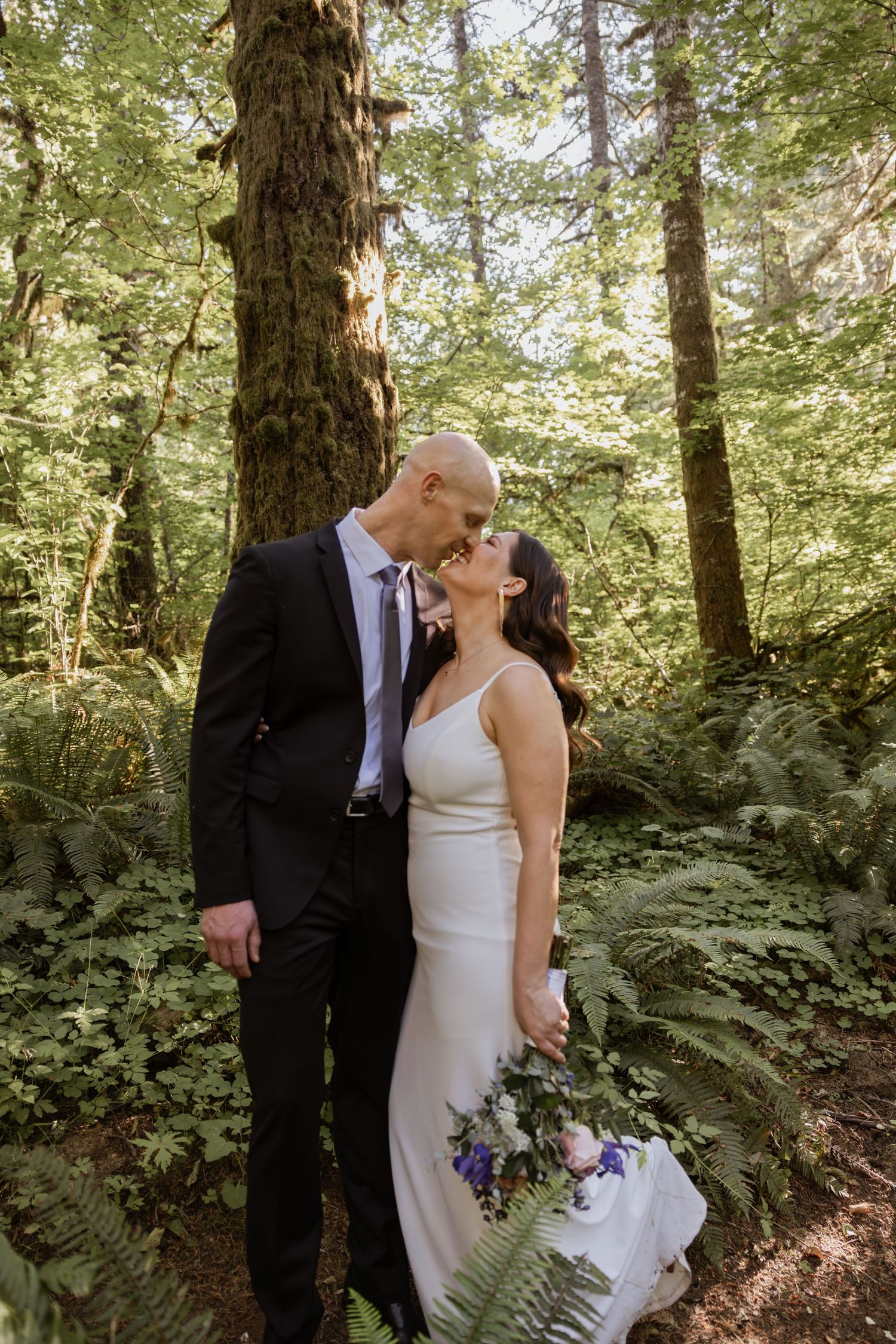 A romantic wedding couple shares an intimate moment in a lush forest setting with tall trees and ferns.