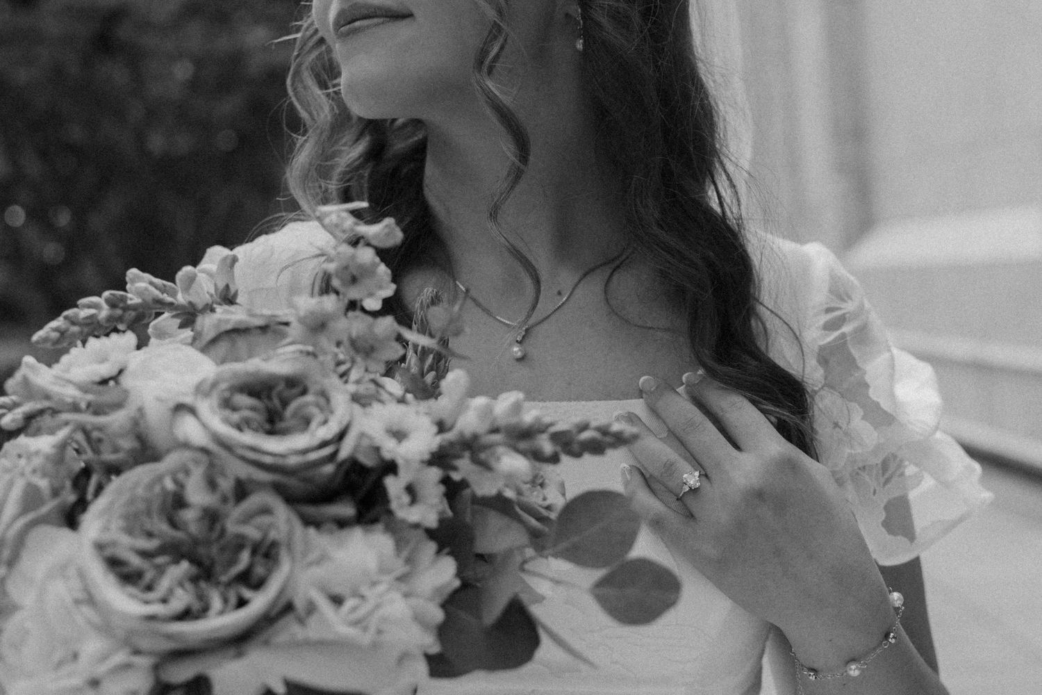 Black and white photo of a bride holding a floral bouquet, wearing a necklace and ring, in a wedding dress.