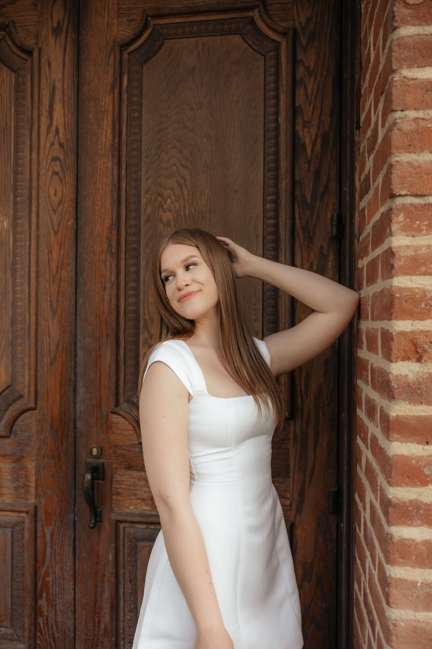 Young woman in white dress poses gracefully by ornate wooden door with brick wall backdrop.