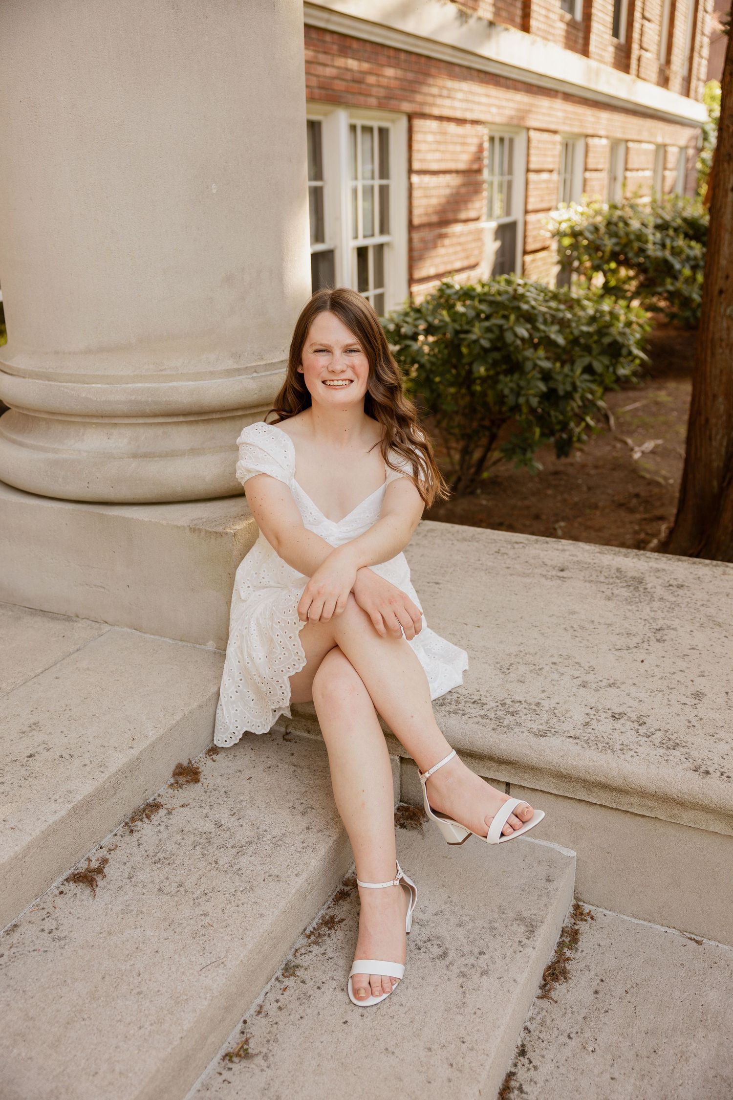 A person in a white dress sits on stone steps by a brick building with white columns and greenery.