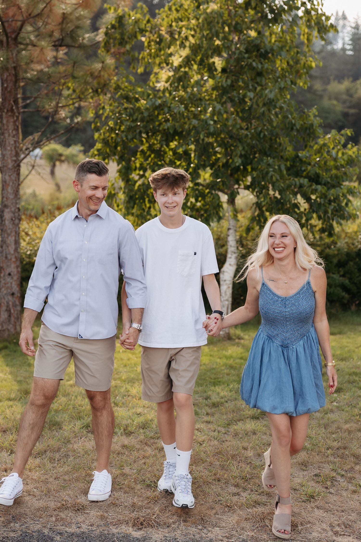 A family in coordinated casual outfits walks together on a grassy hill during a sunny afternoon photoshoot.
