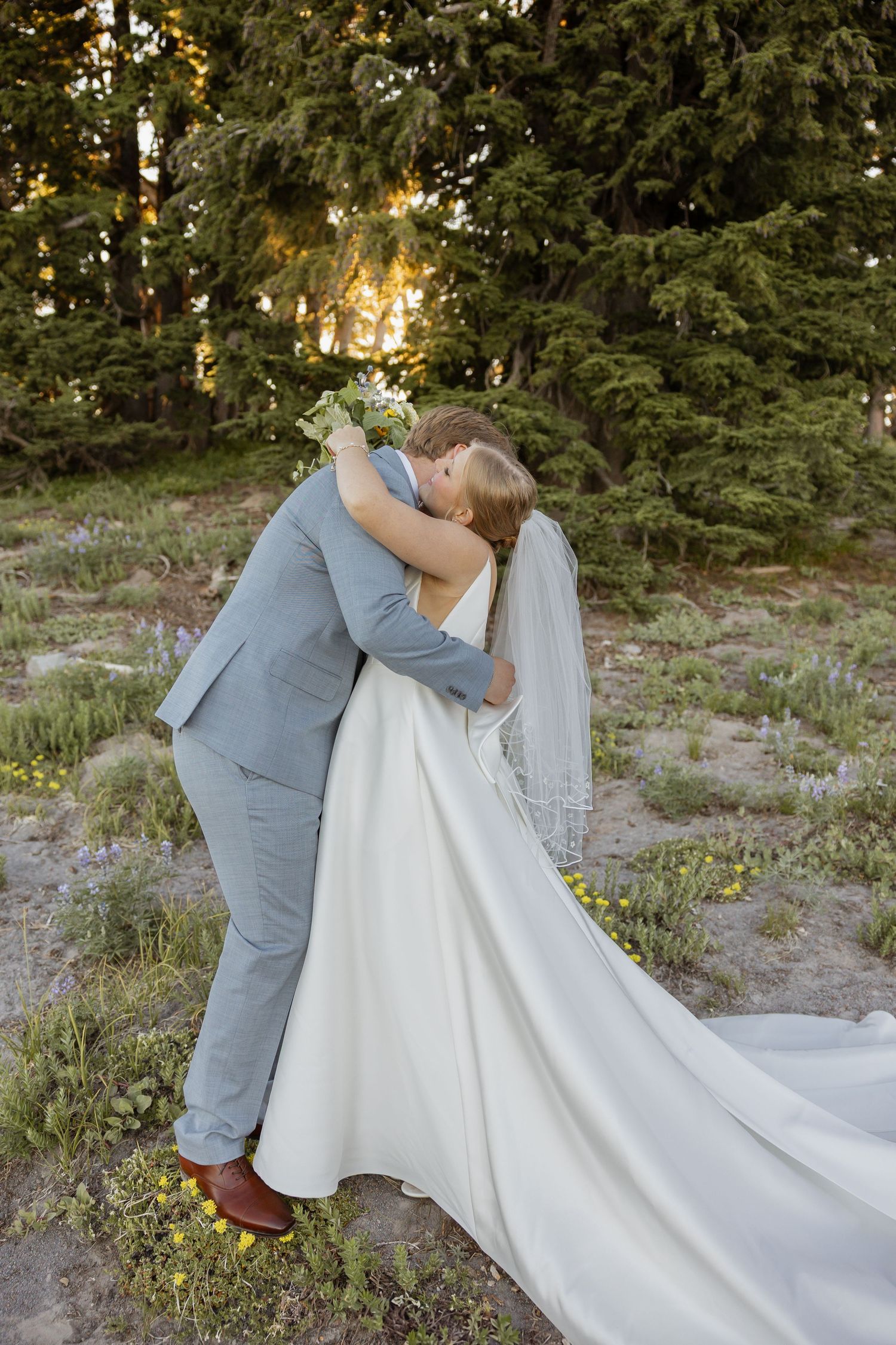 A couple in wedding attire share a romantic kiss outdoors surrounded by evergreen trees at sunset.