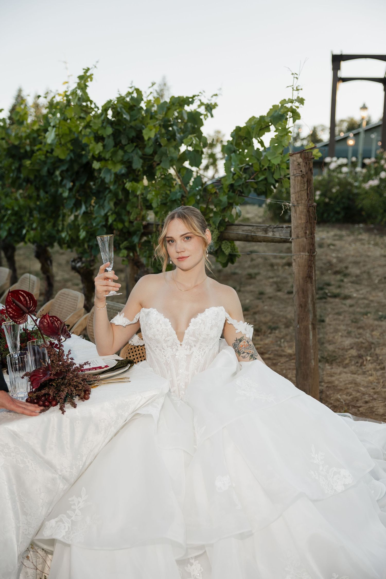Bride in white lace ball gown raising champagne glass at vineyard table with lush green vines backdrop.