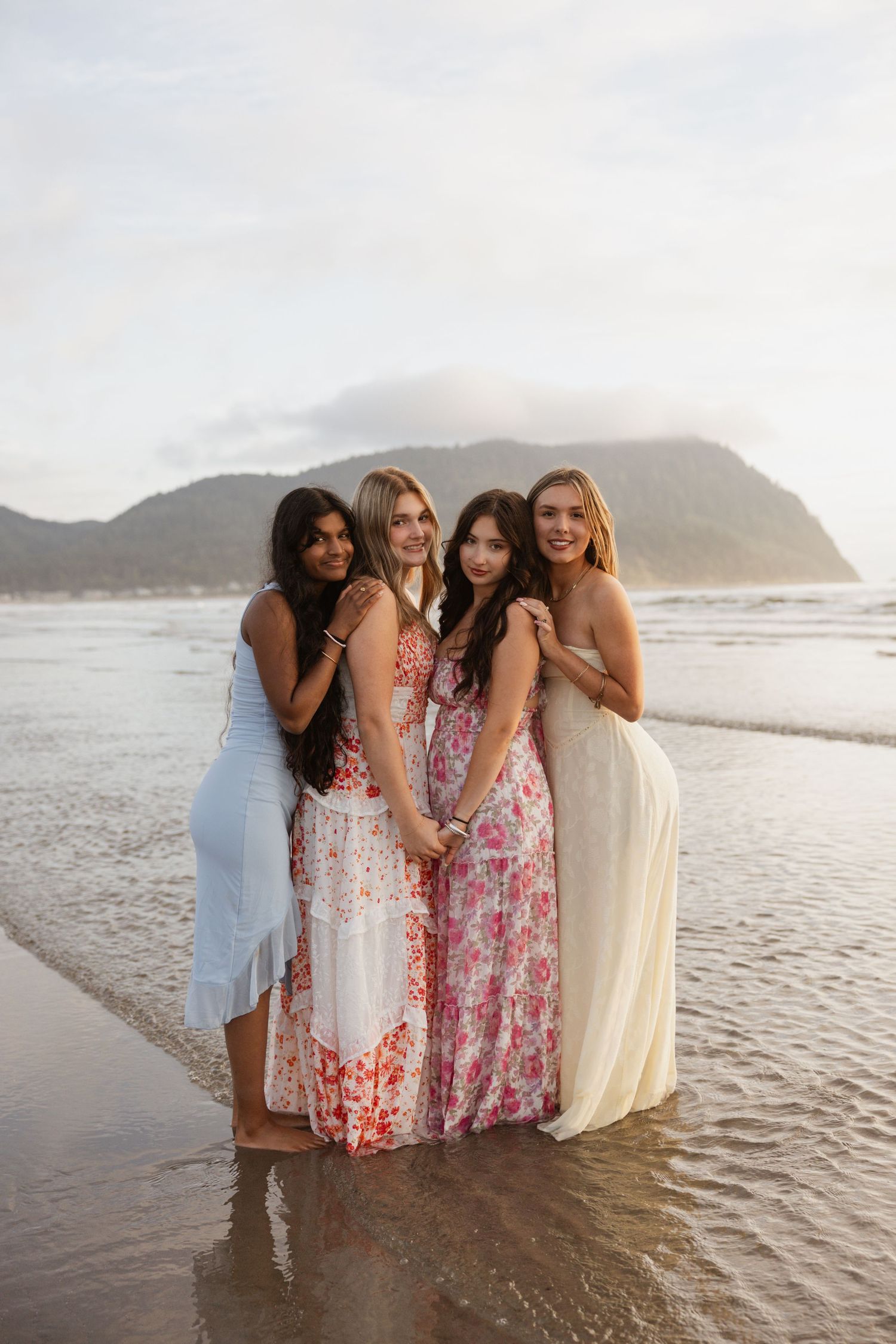 A group of friends in floral dresses pose together on a beach at sunset with mountains in the background.
