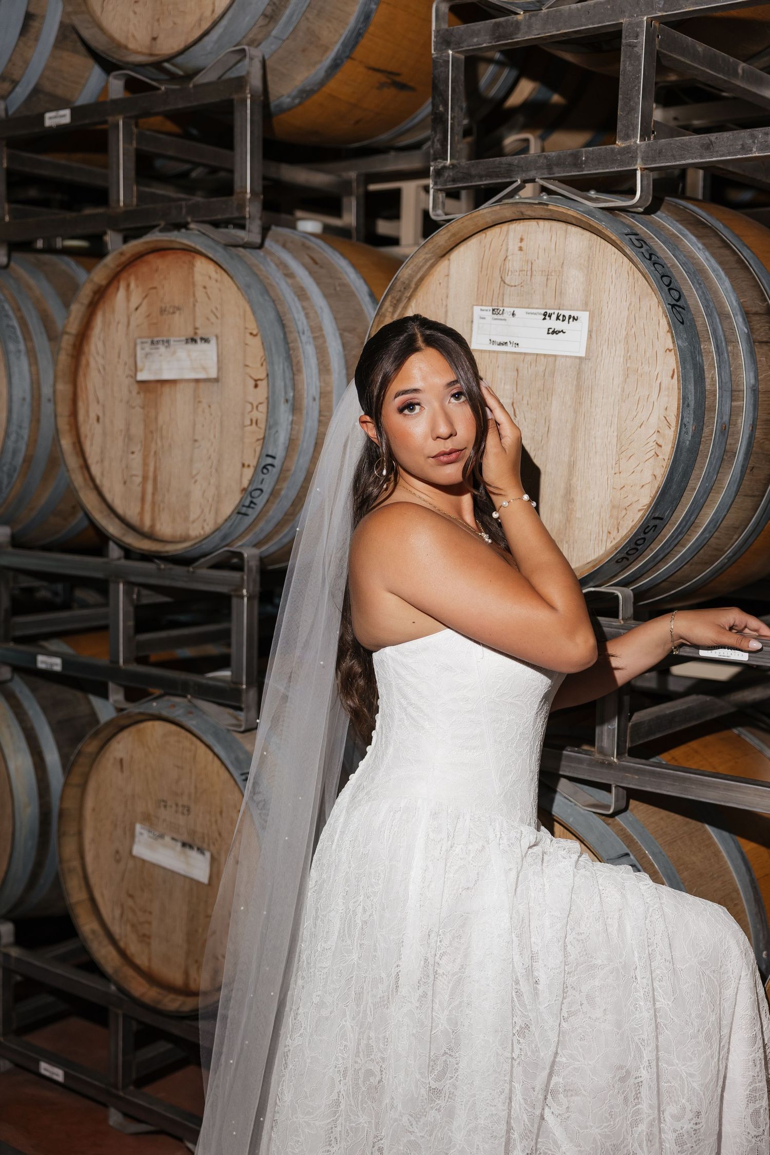 Bride in white strapless gown and veil posing elegantly among oak wine barrels in a winery cellar.