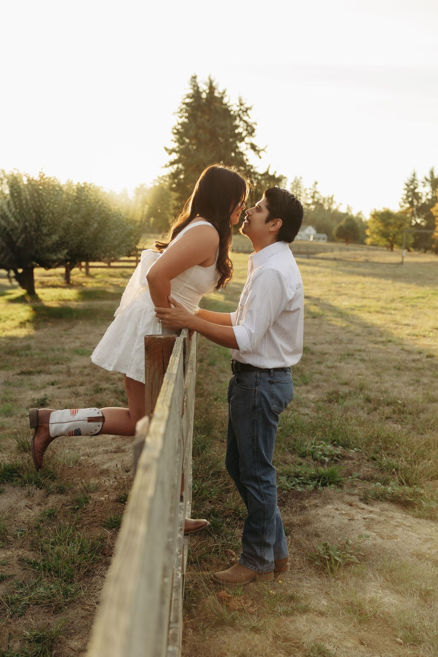 Romantic couple in white outfits leaning against wooden fence during sunset photo session.