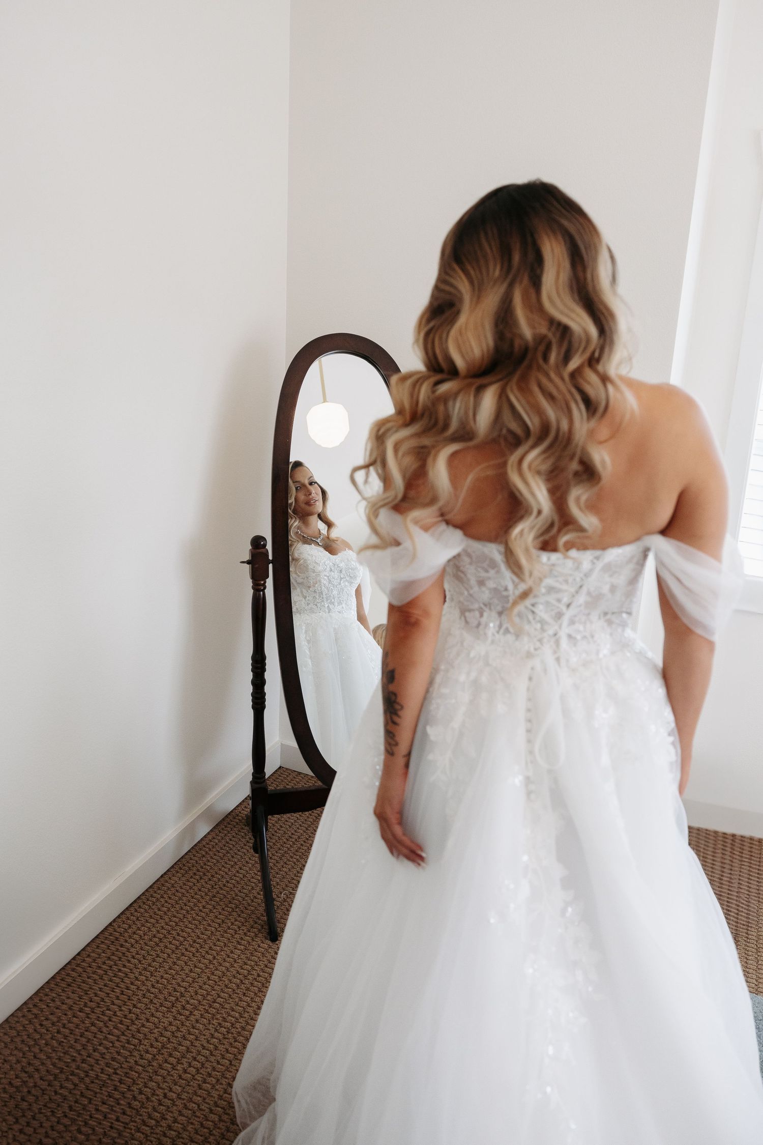 Back view of a bride in a lace wedding gown looking in a full-length mirror.