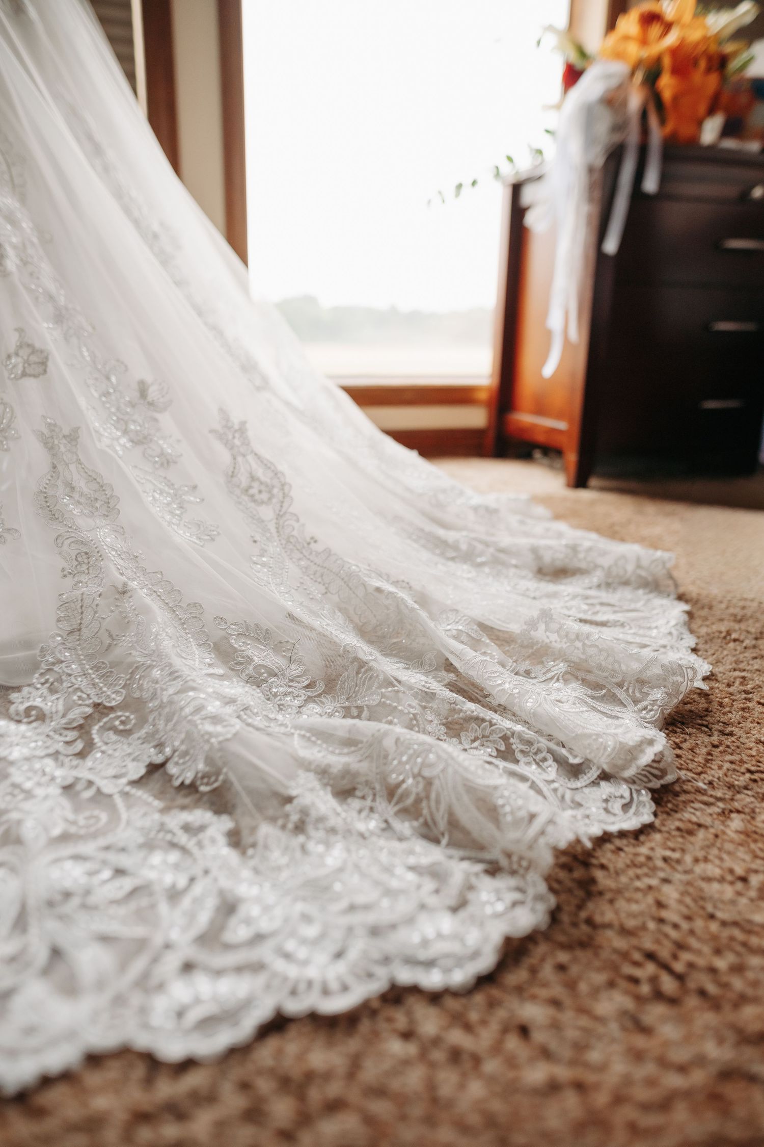 Delicate lace details along the hem of a white wedding dress train on beige carpet.