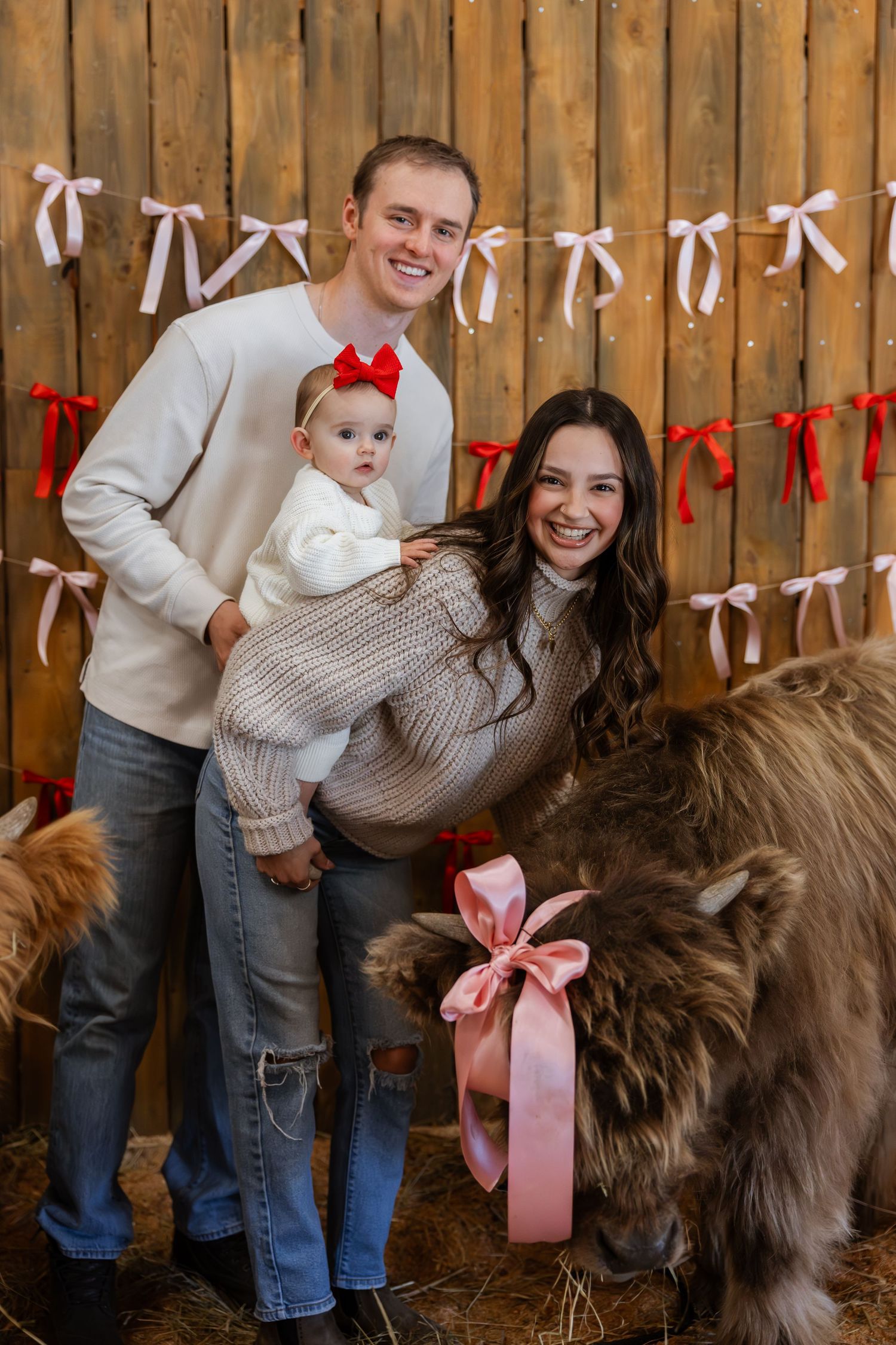 Family poses with their large brown dog against wooden fence decorated with white and red ribbons for holiday photos.