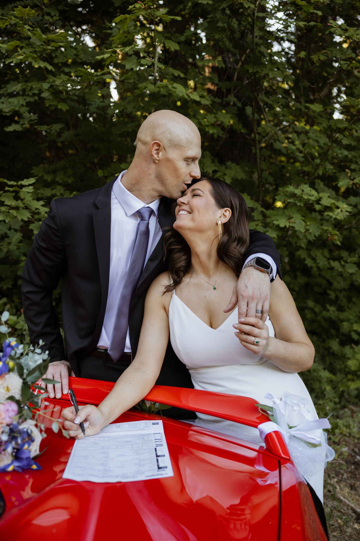 A couple in wedding attire share a tender moment while leaning against a bright red sports car.