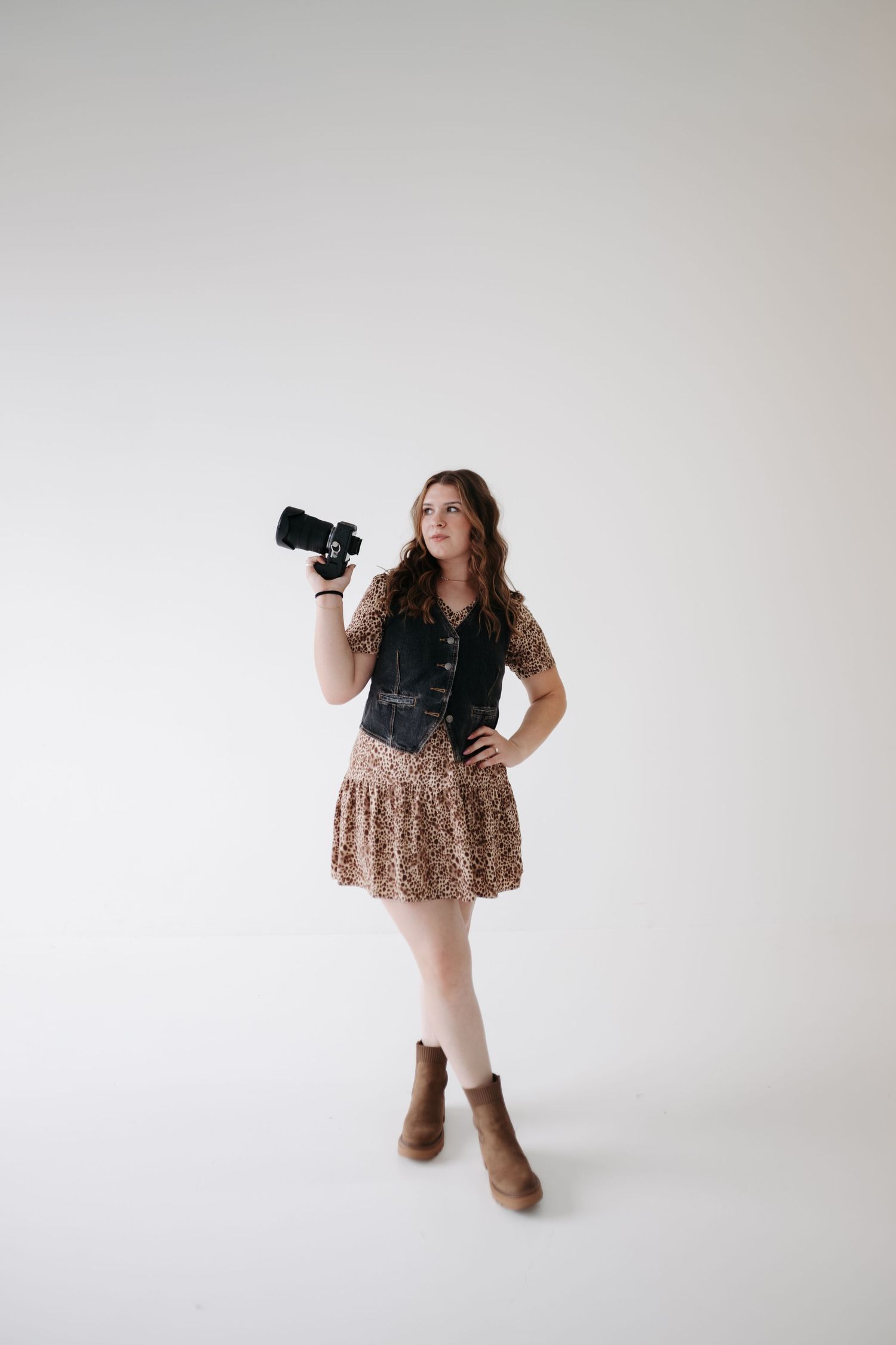 Professional photographer in a leopard print dress and black vest poses with camera against white backdrop.
