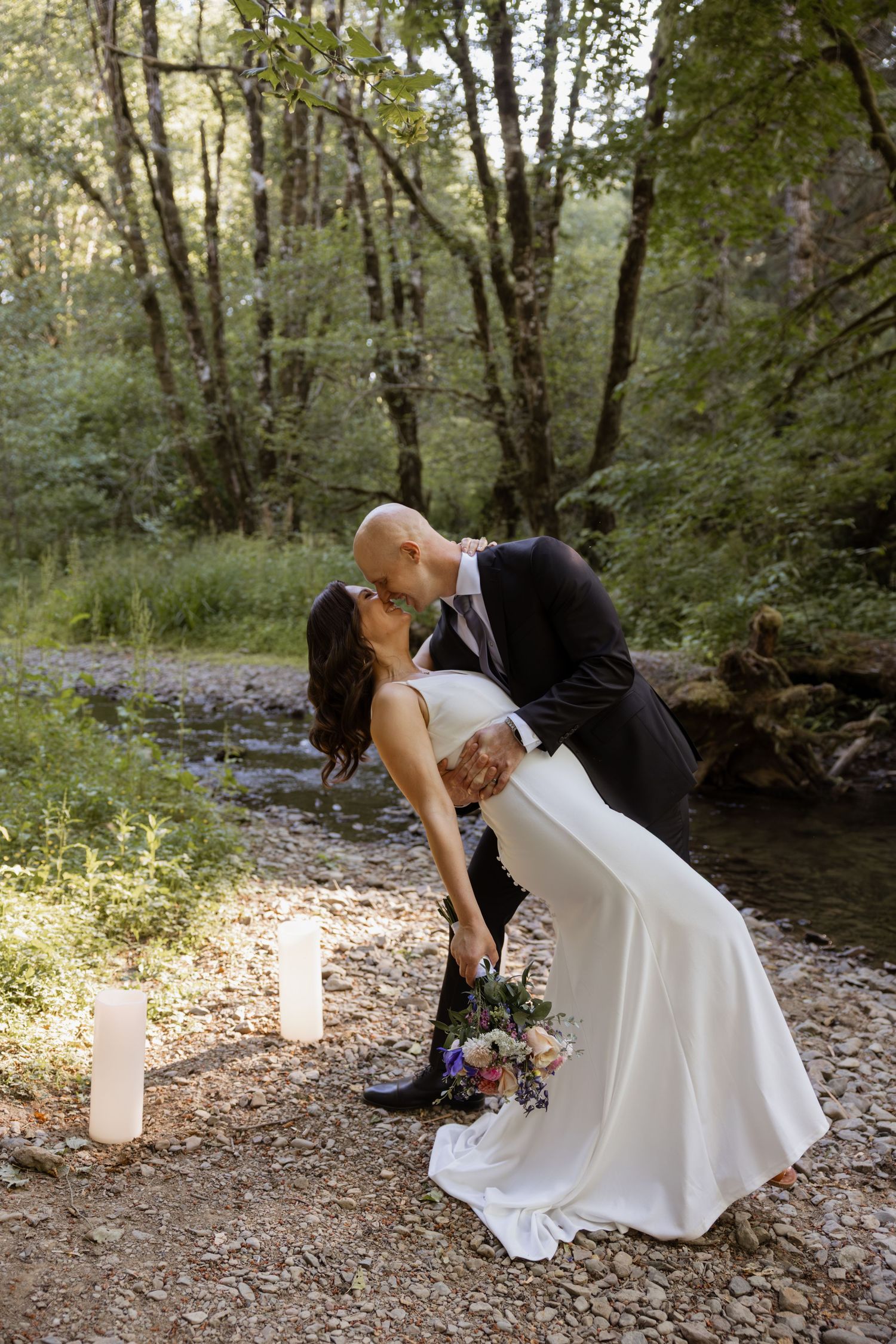 A couple in wedding attire performs a romantic dip pose in a wooded outdoor setting with glowing candles.