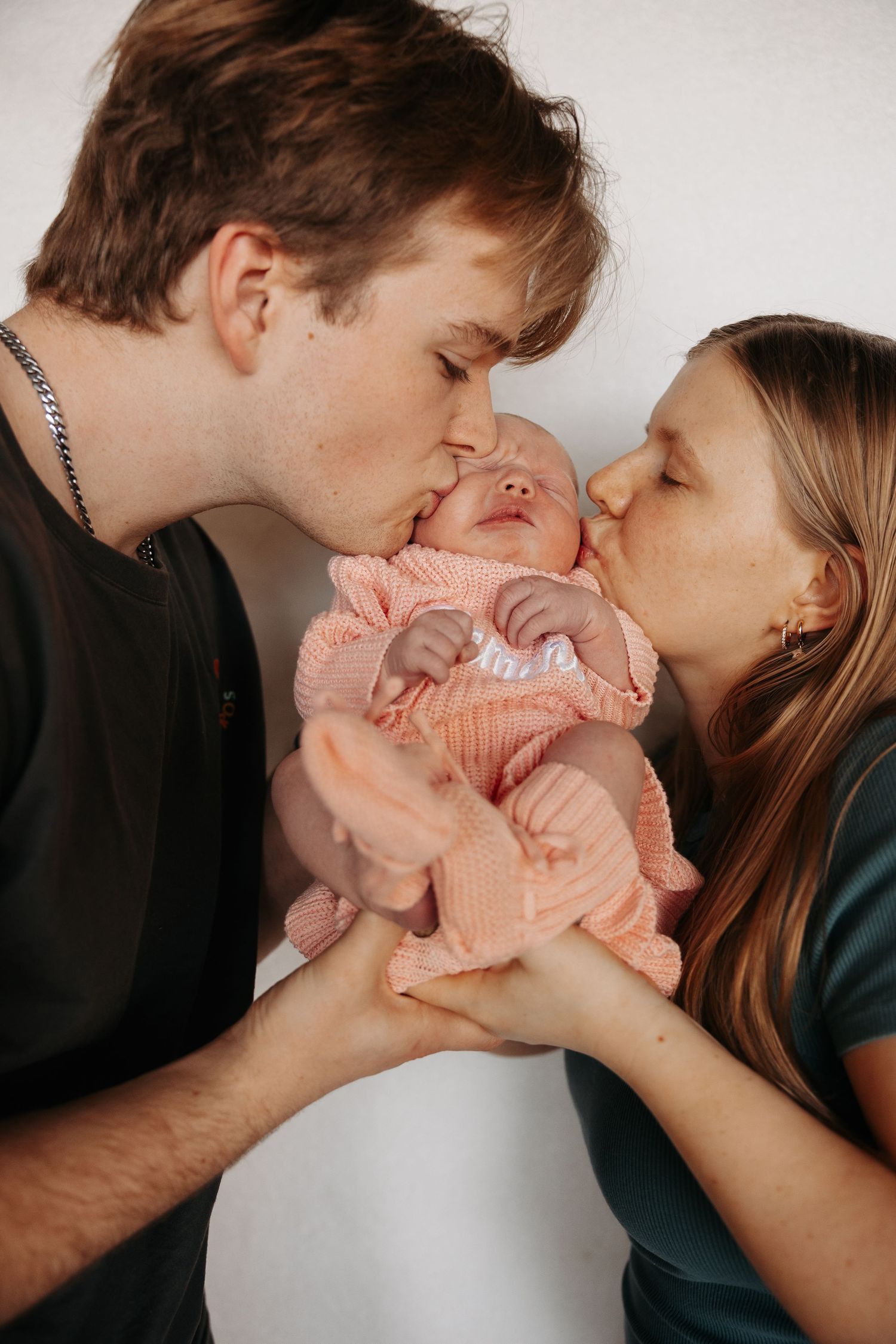 A couple shares tender kisses while holding their newborn baby wrapped in a pink blanket.